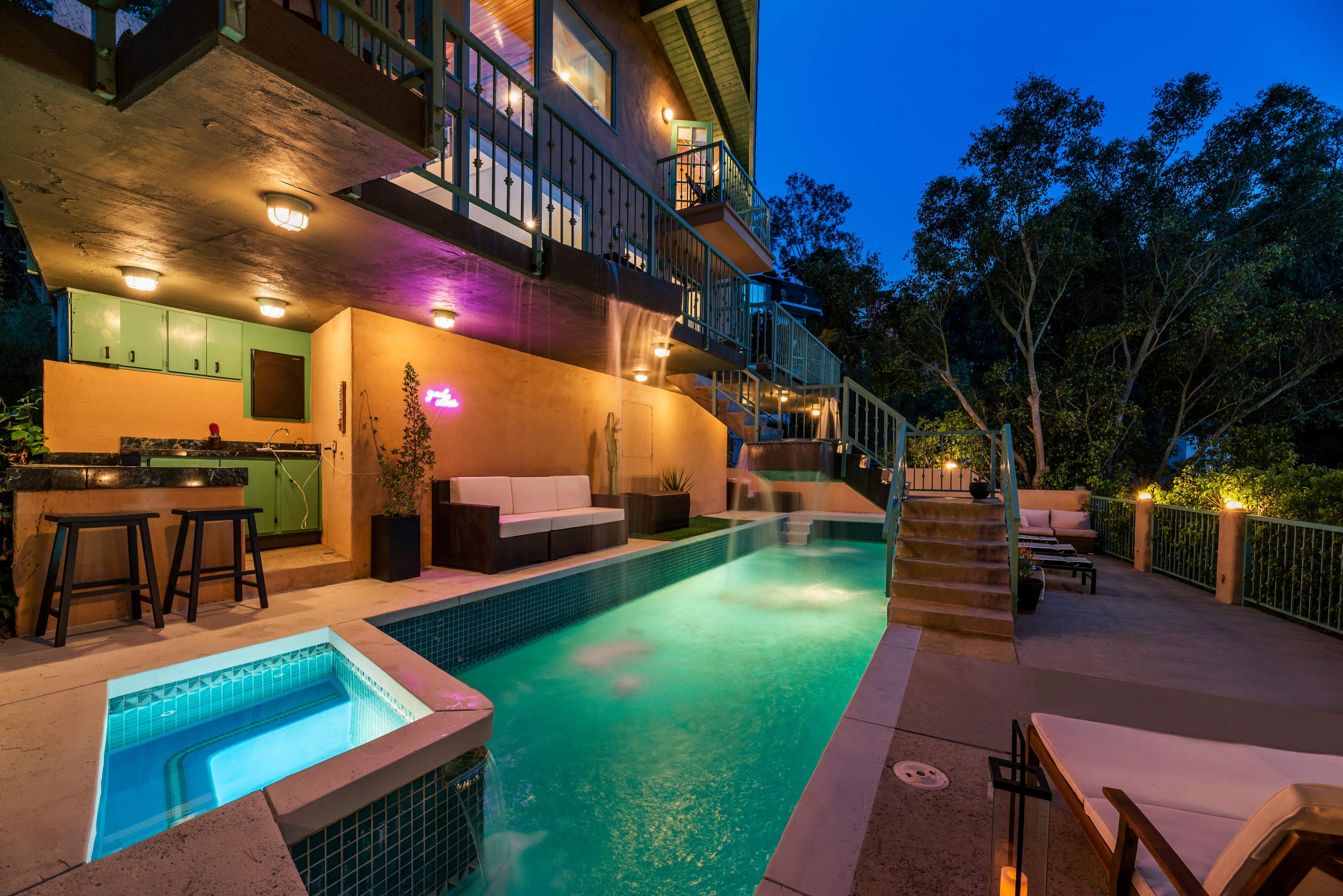 A modern home with a swimming pool and spa is illuminated at dusk, surrounded by trees and patio seating.