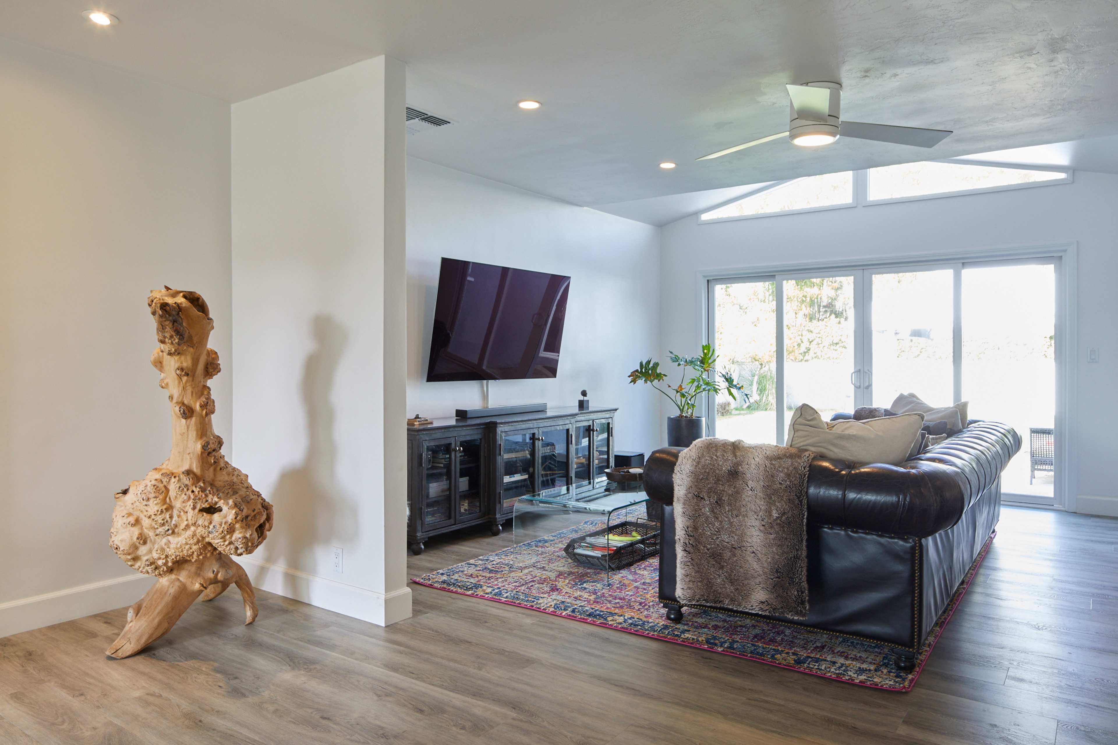 The image shows a modern living room featuring a leather sofa, a large television mounted on the wall, and a decorative wooden sculpture in a bright, airy space.