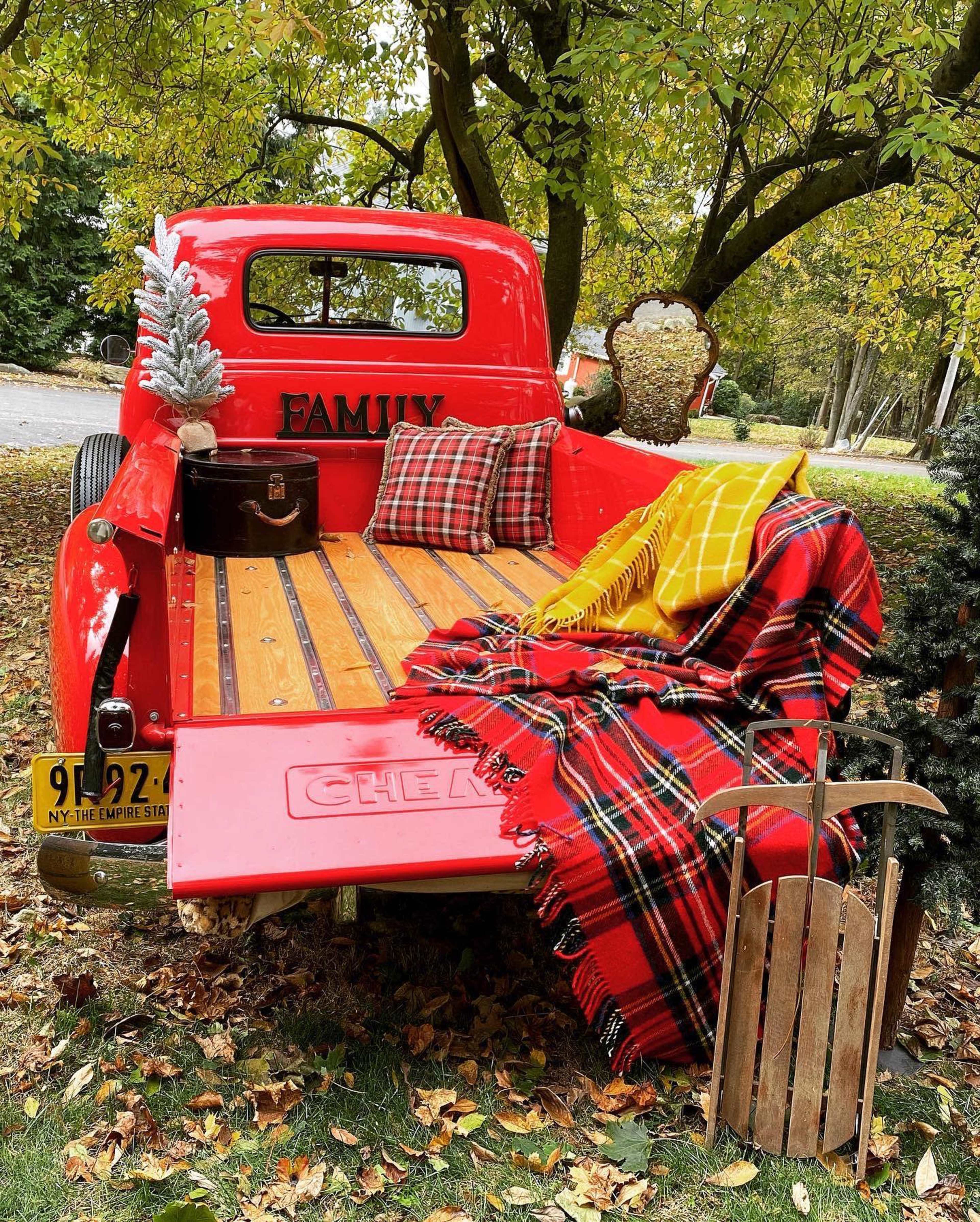 A vintage red pickup truck is parked under trees, featuring a plaid blanket, pillows, a wooden crate, and decorative items in the truck bed.