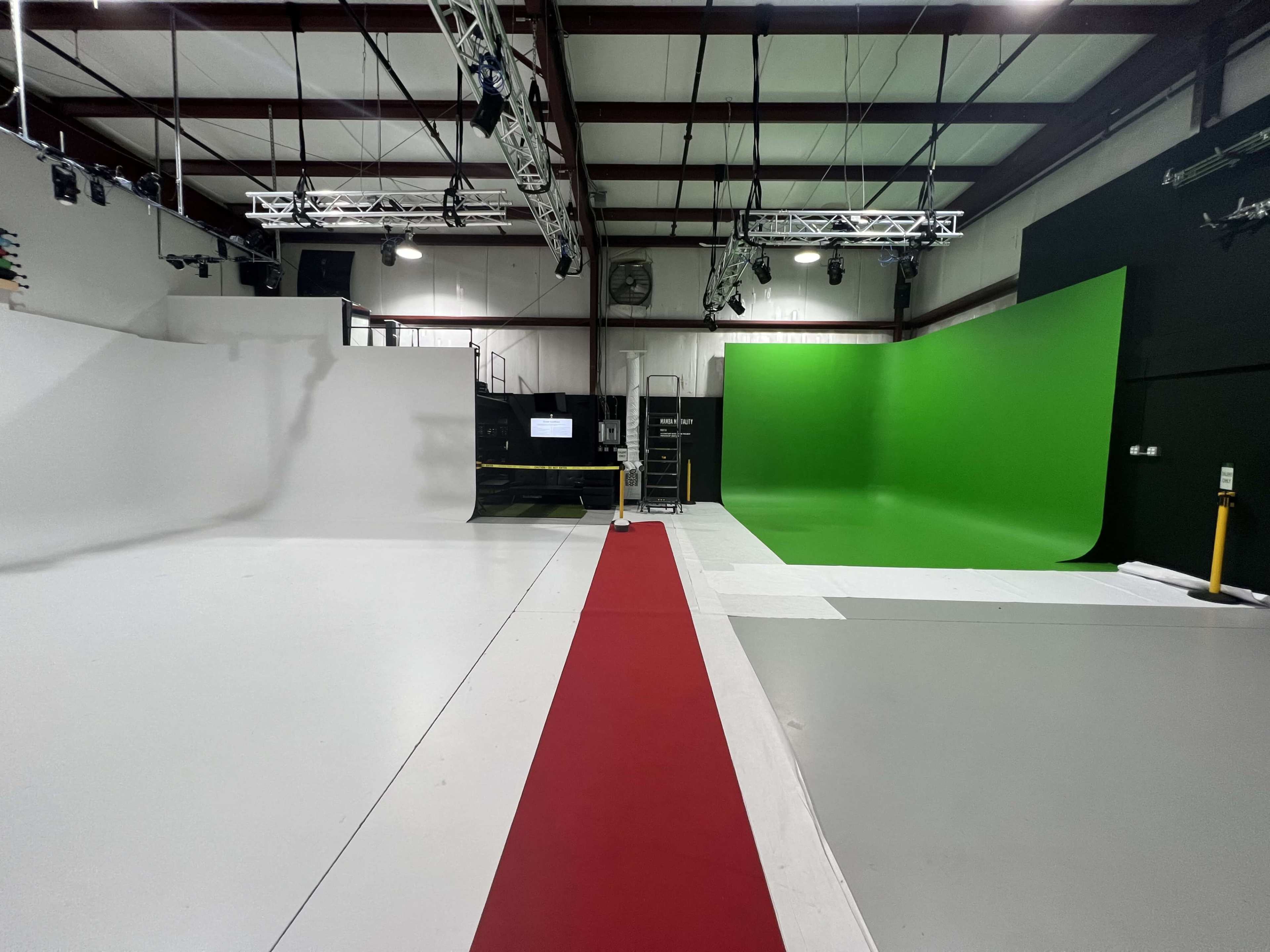 The image shows a production studio featuring a red carpet leading to white and green studio backdrops, with various lighting fixtures mounted above.