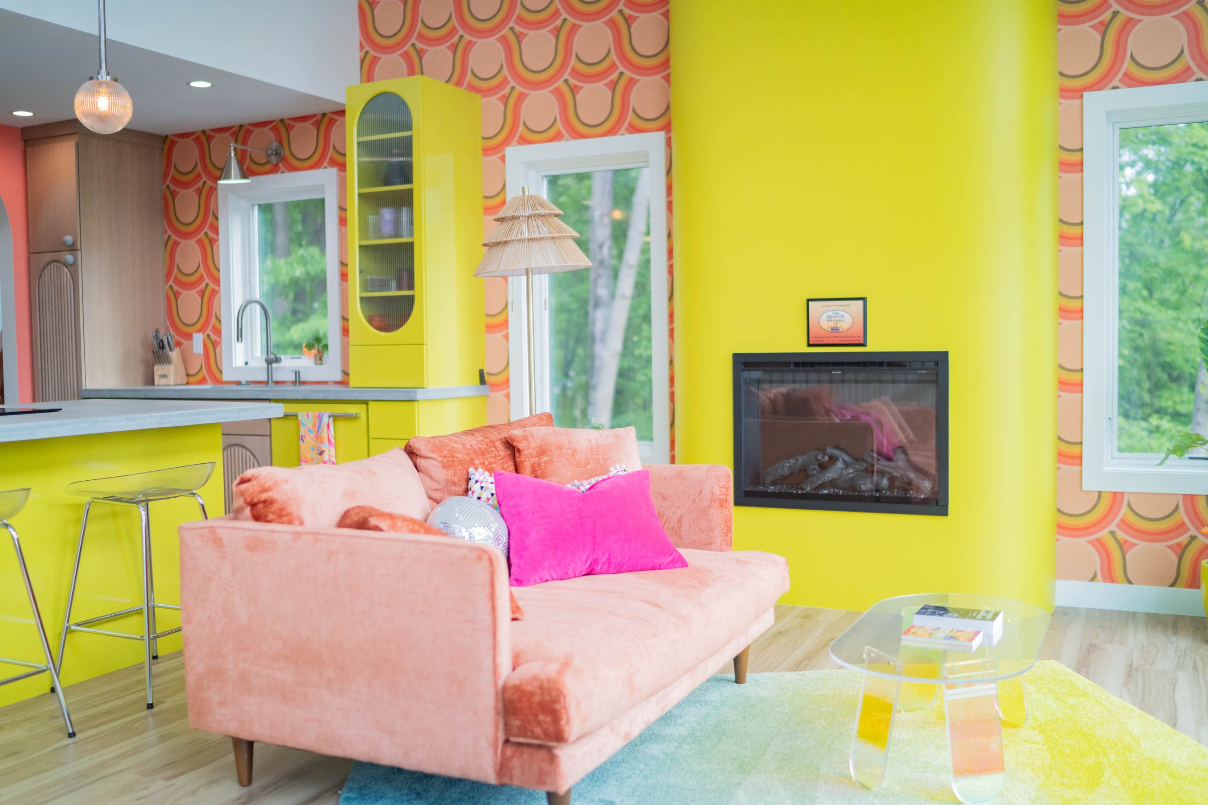 The image shows a modern living room with a bright yellow wall, an orange sofa, and a contemporary fireplace beside a kitchen area featuring a sleek counter.