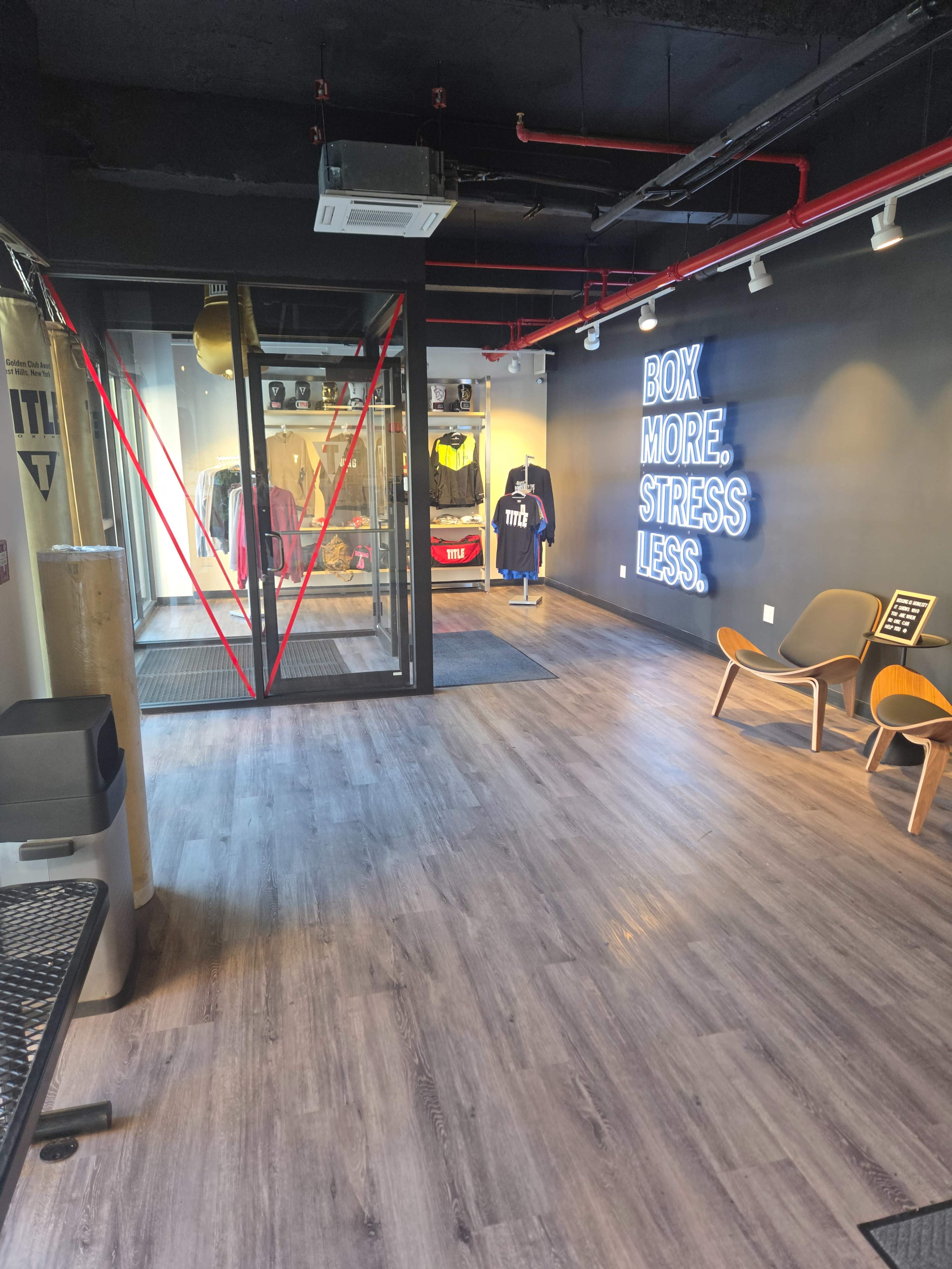 The image shows a modern interior of a retail space, featuring wooden flooring, lounge chairs, and a neon sign that reads "BOX MORE.. STRESS LESS," along with glass doors displaying apparel.