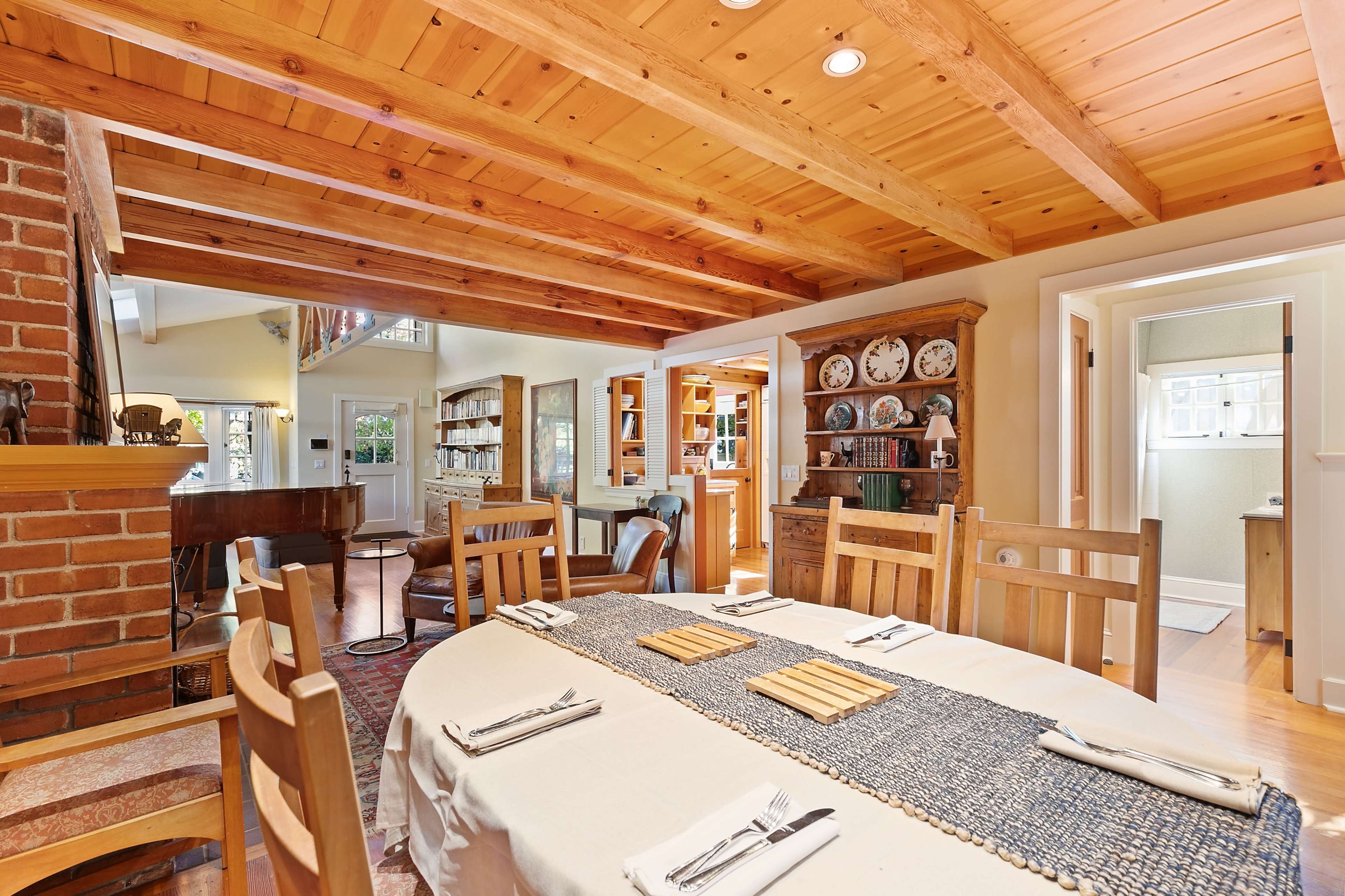 A dining area features a wooden table set with utensils and a placemat, surrounded by chairs and illuminated by natural light from nearby windows.