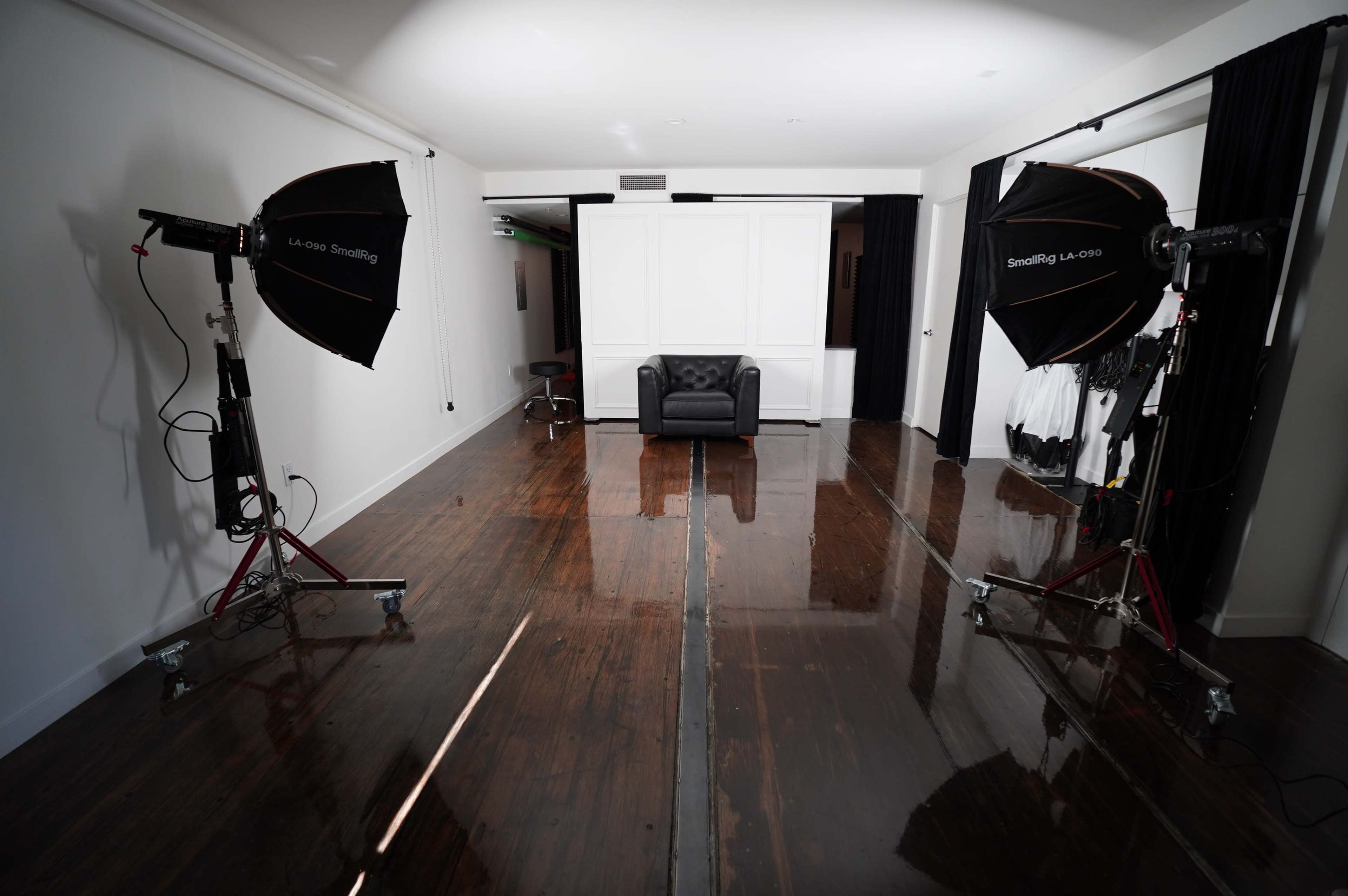 The image shows a photography studio featuring two large softbox lights positioned on either side of a black leather couch in the center of a reflective wooden floor.
