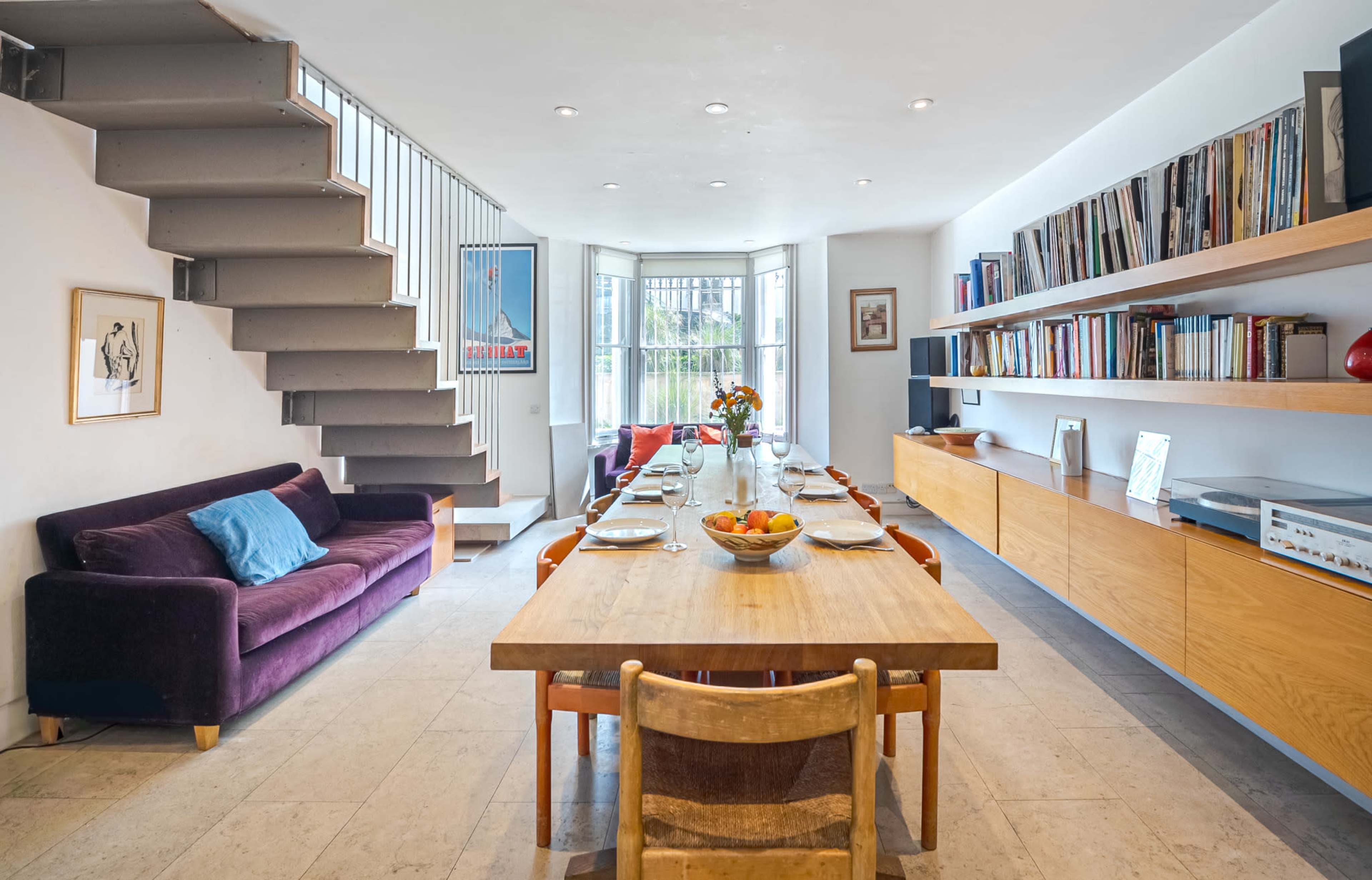 A dining area features a wooden table set for eight, surrounded by chairs, with a sofa and bookshelves along the walls and stairs leading to the upper level.