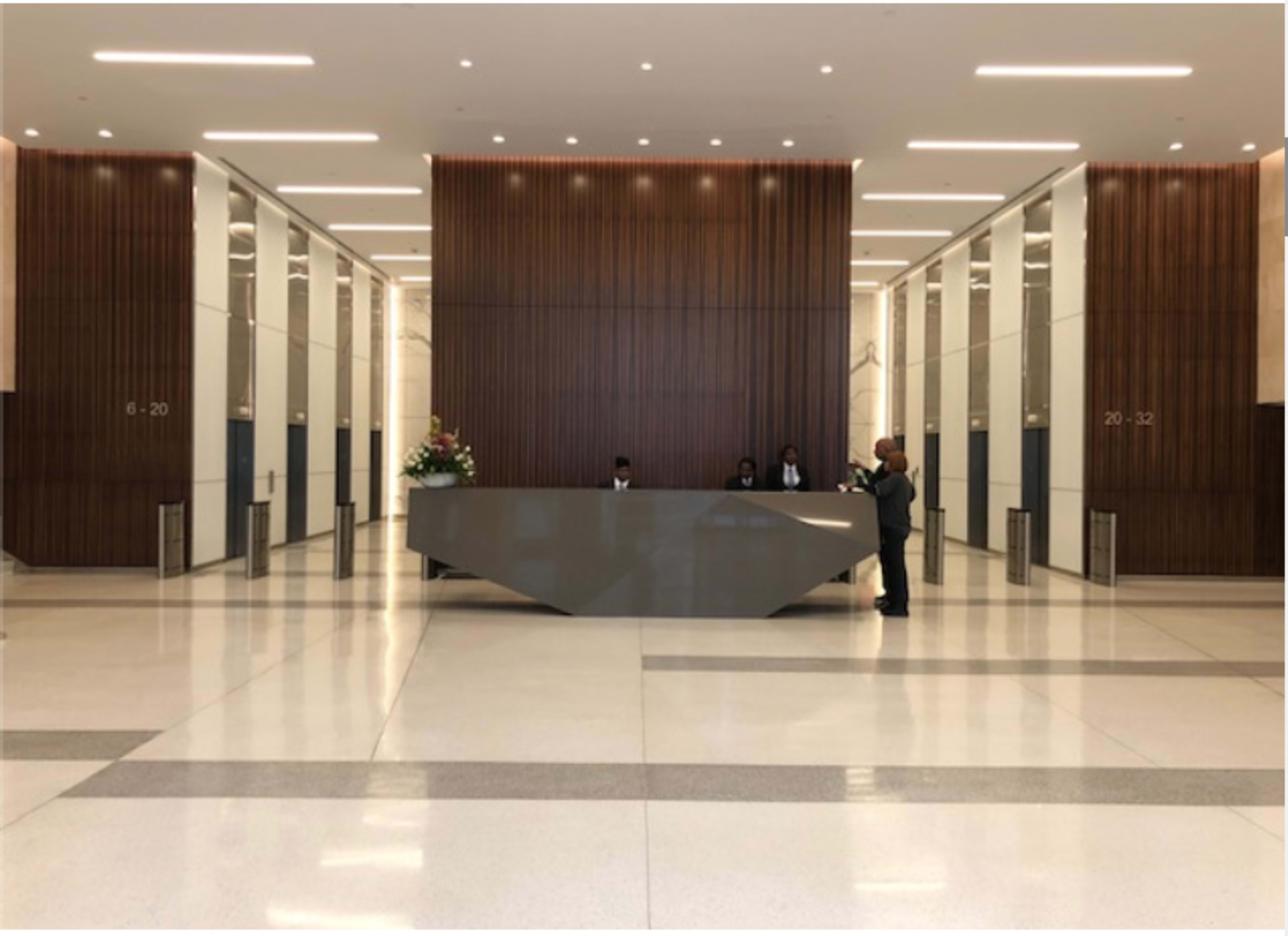 The image shows a modern lobby area with a large reception desk and three receptionists in front of a wooden wall and polished floors.