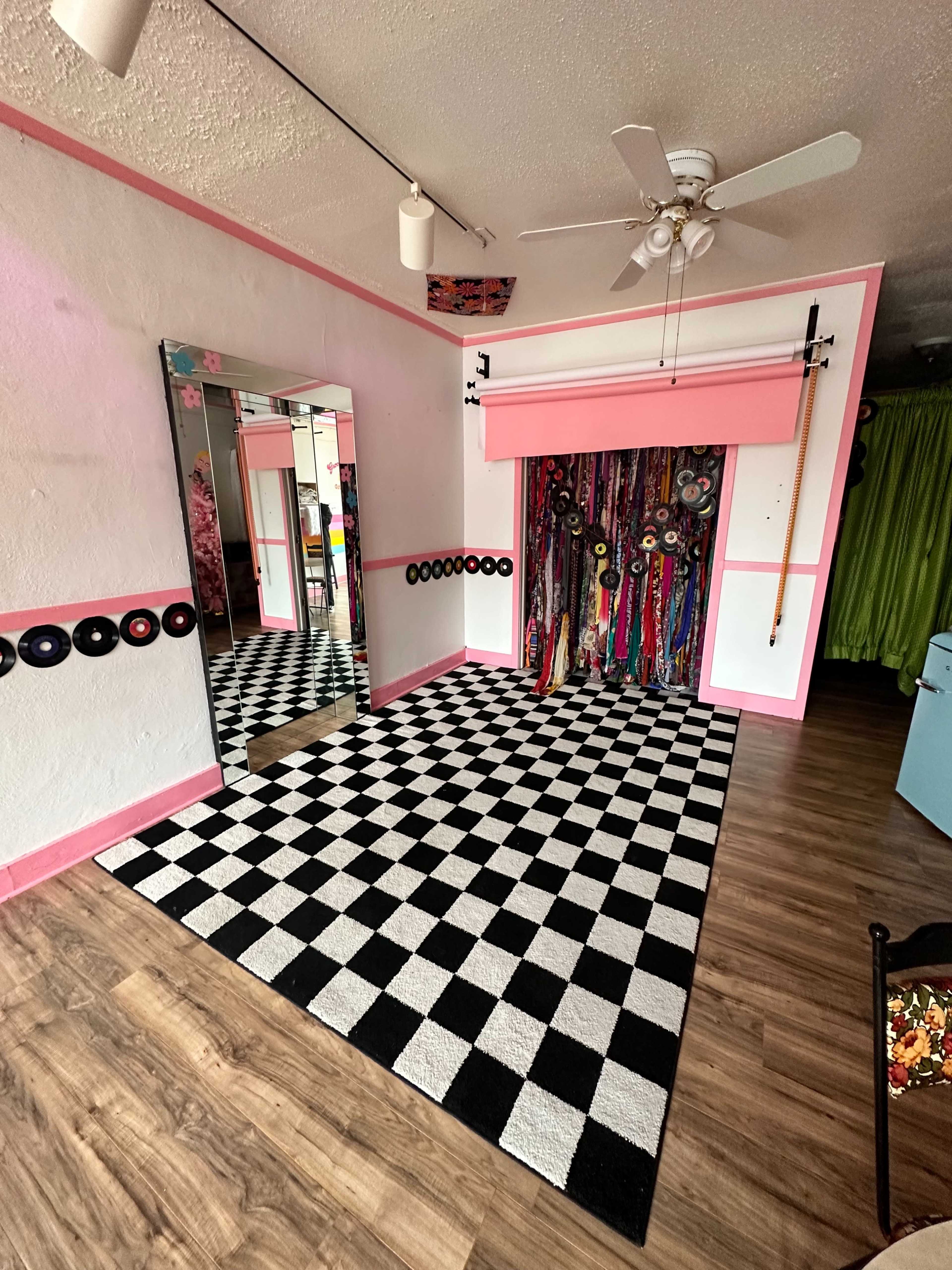 The room features a checkered black-and-white floor, a large mirror, and a colorful wall of fabric drapes, with pink accents throughout.