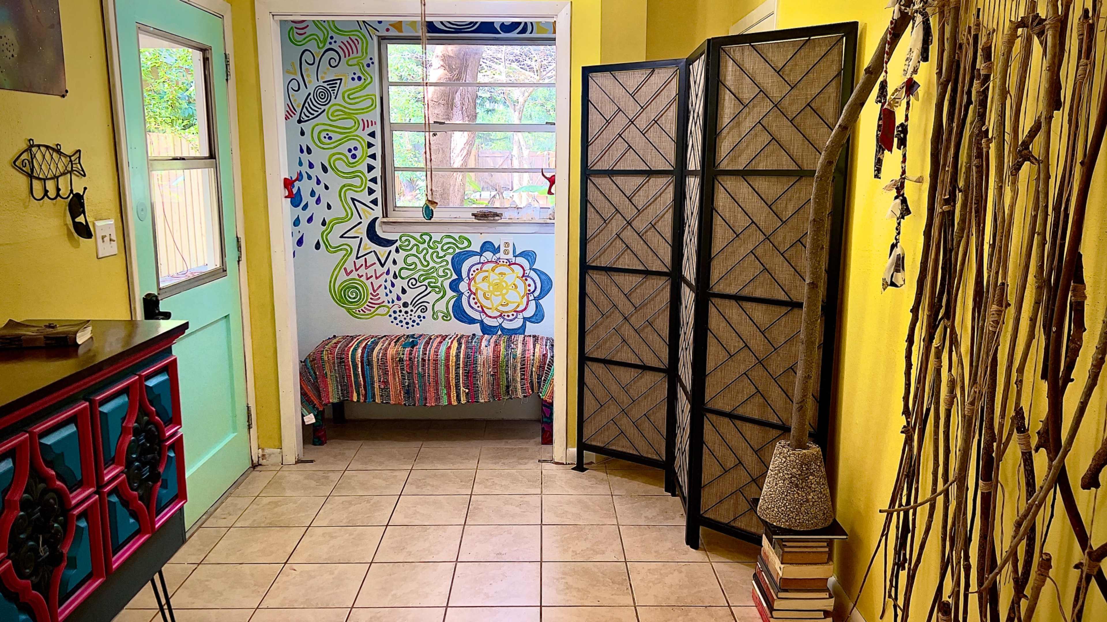 The image shows a brightly colored entryway featuring a decorative wall mural, a bench, a folding screen, and a stack of books beside a tall decorative piece.