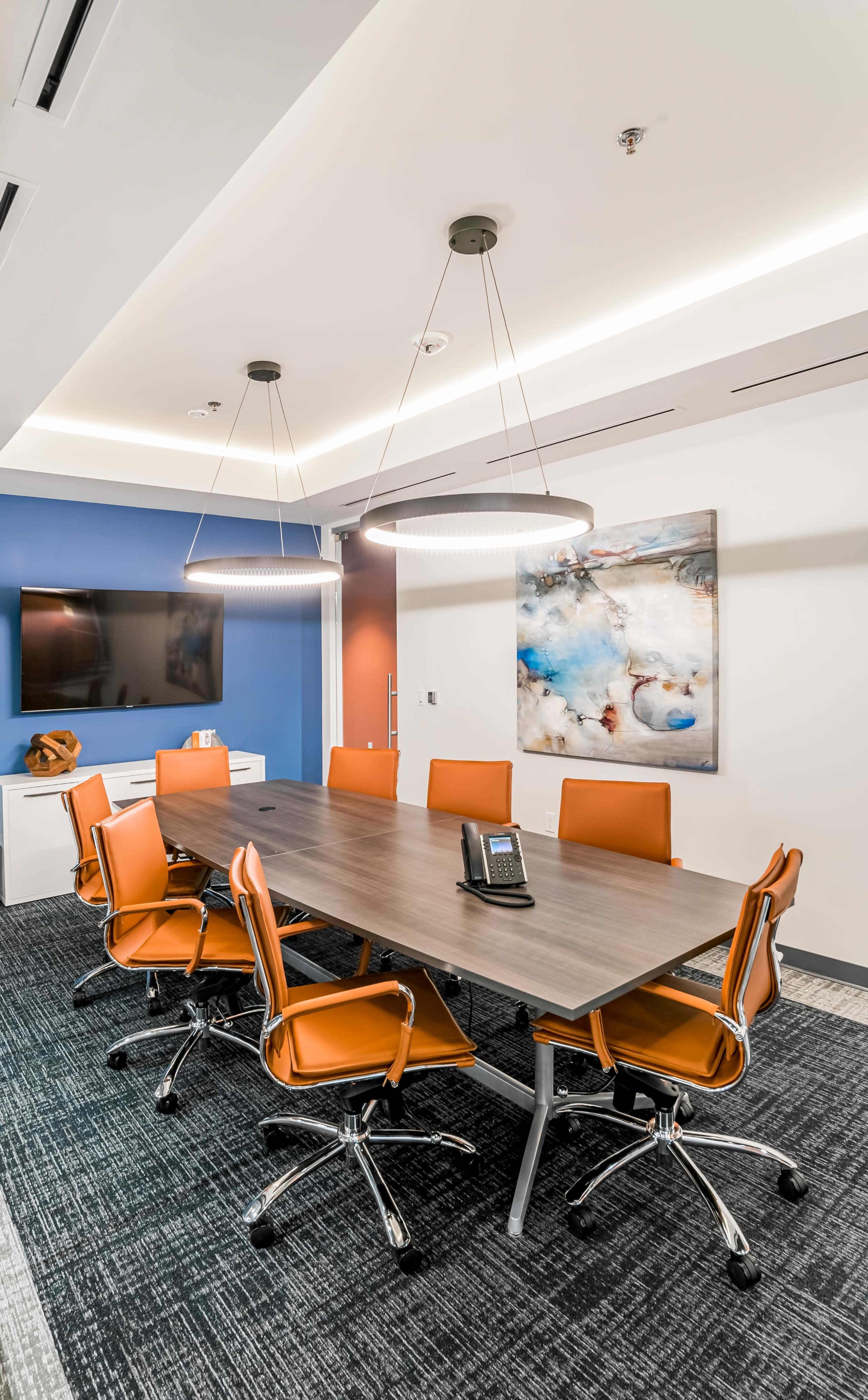 A modern conference room features a long wooden table surrounded by orange chairs, with dimmable pendant lights above and a TV mounted on the wall.