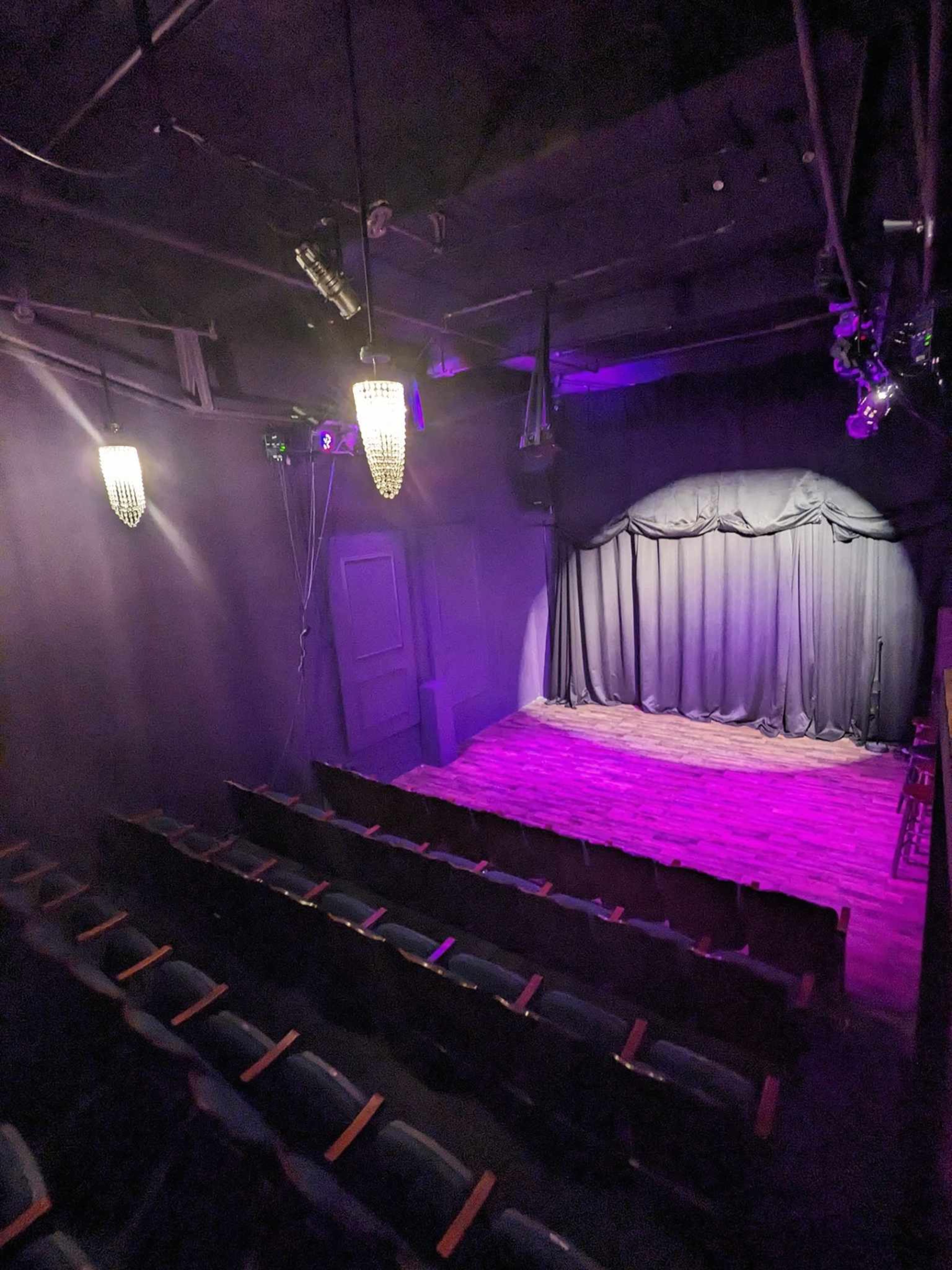 An empty theater stage with a dark curtain and rows of green seats facing it, illuminated by purple lights.