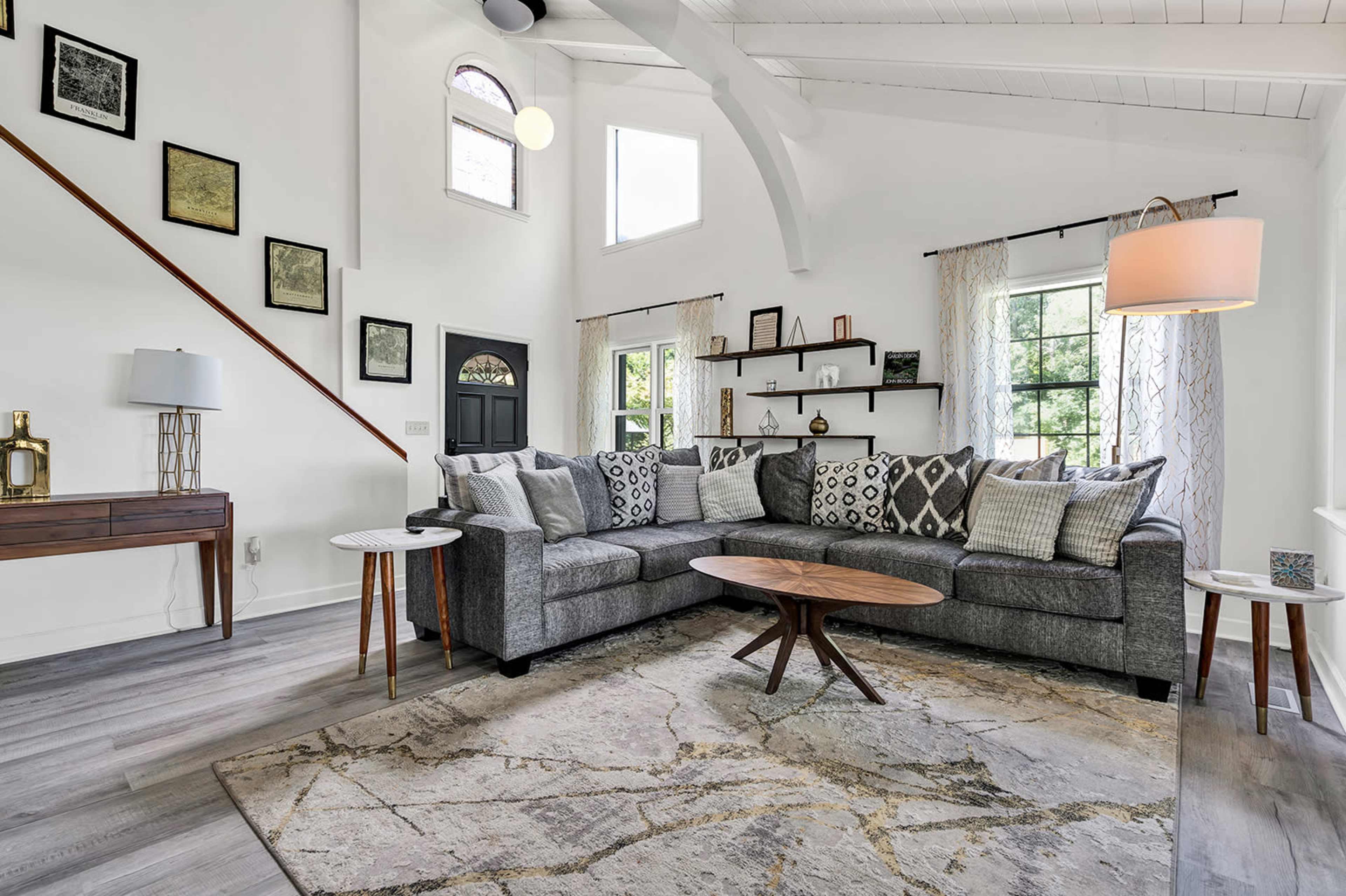 The image shows a bright living room with a large sectional sofa, a coffee table, and decorative shelves against the walls.