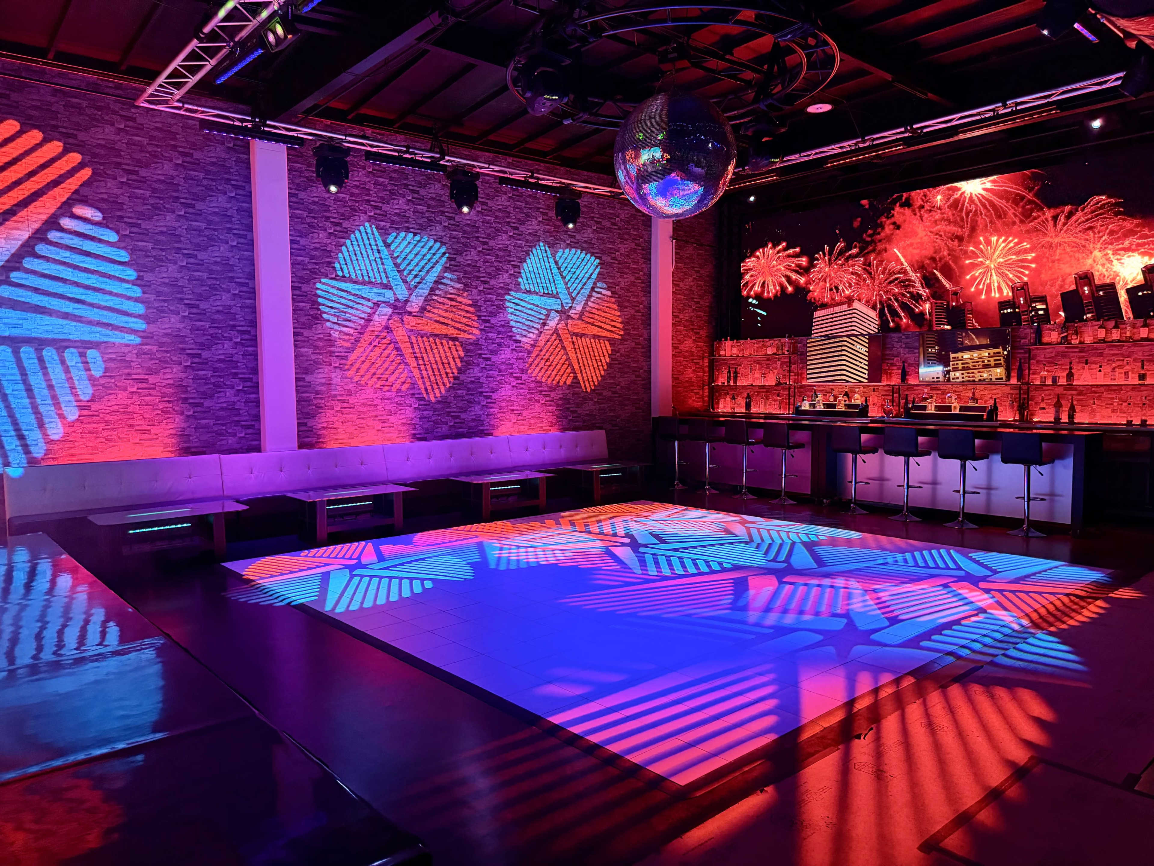 The image shows a vibrant nightclub interior with colorful lighting effects on the walls and dance floor, along with a bar area in the background.