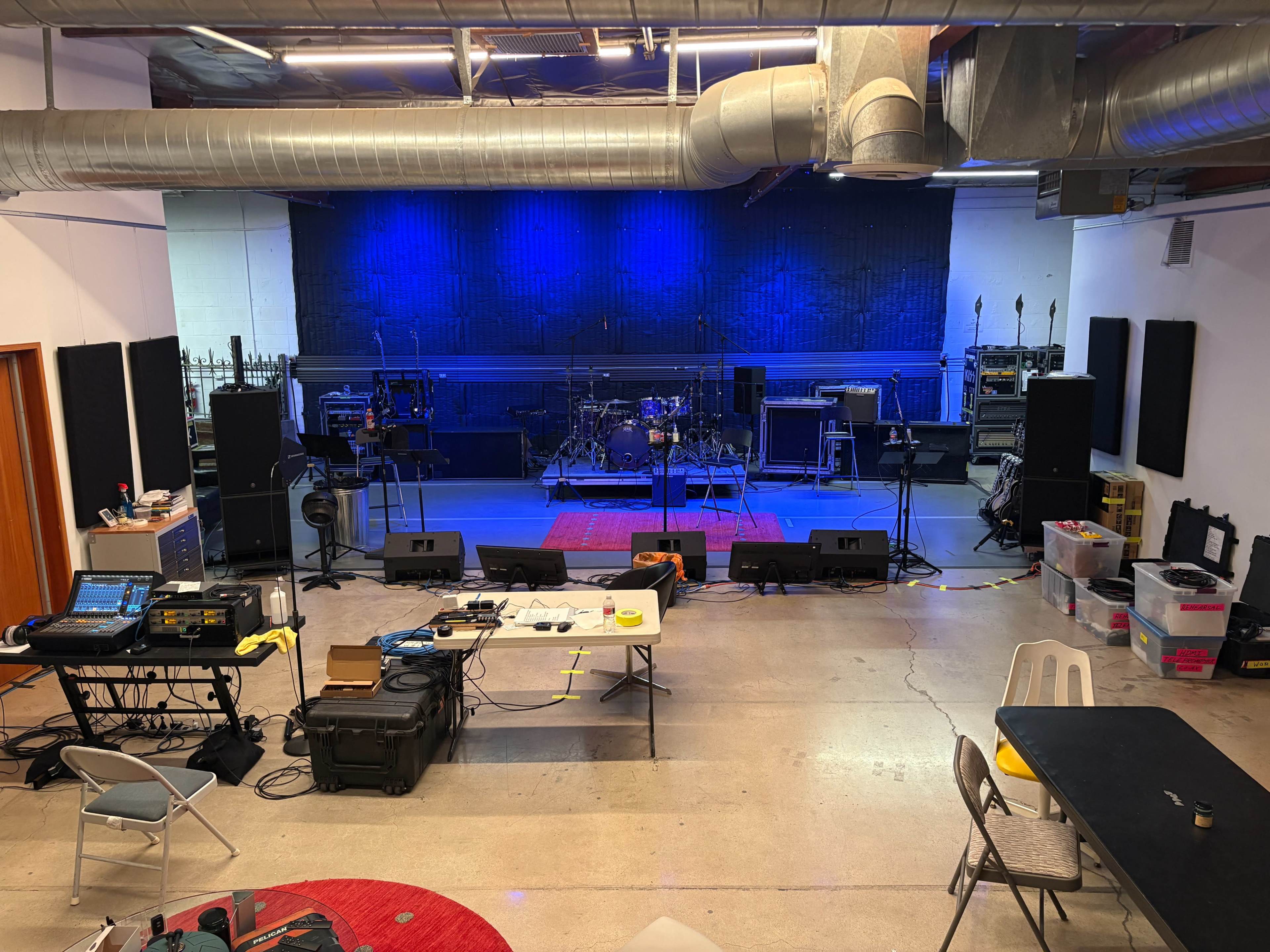 The image shows a music rehearsal space with instruments, amplifiers, and sound equipment arranged on a stage, illuminated by blue lighting.