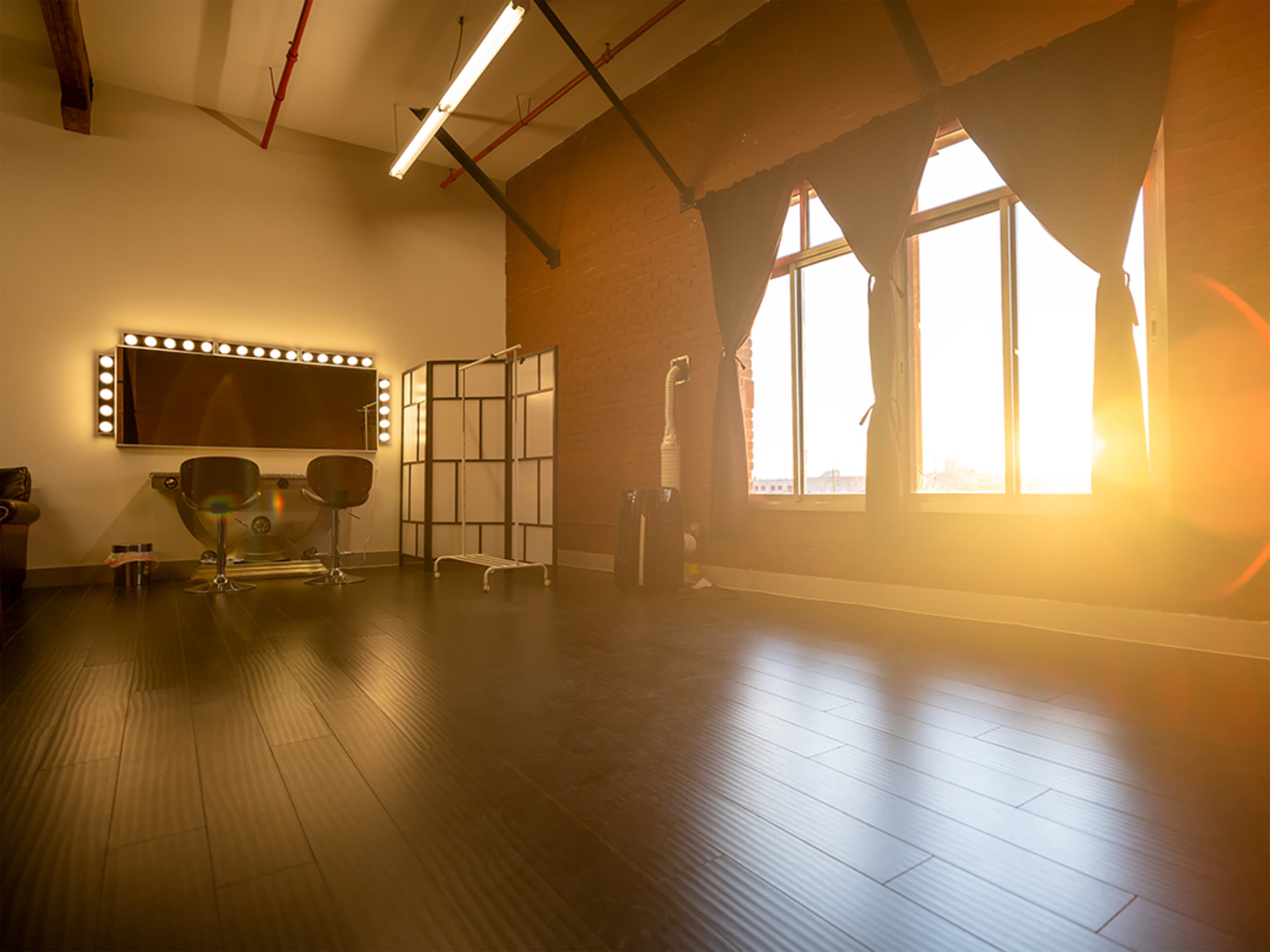 A spacious room with a mirrored vanity, two black chairs, a folding screen, and large windows letting in sunlight.