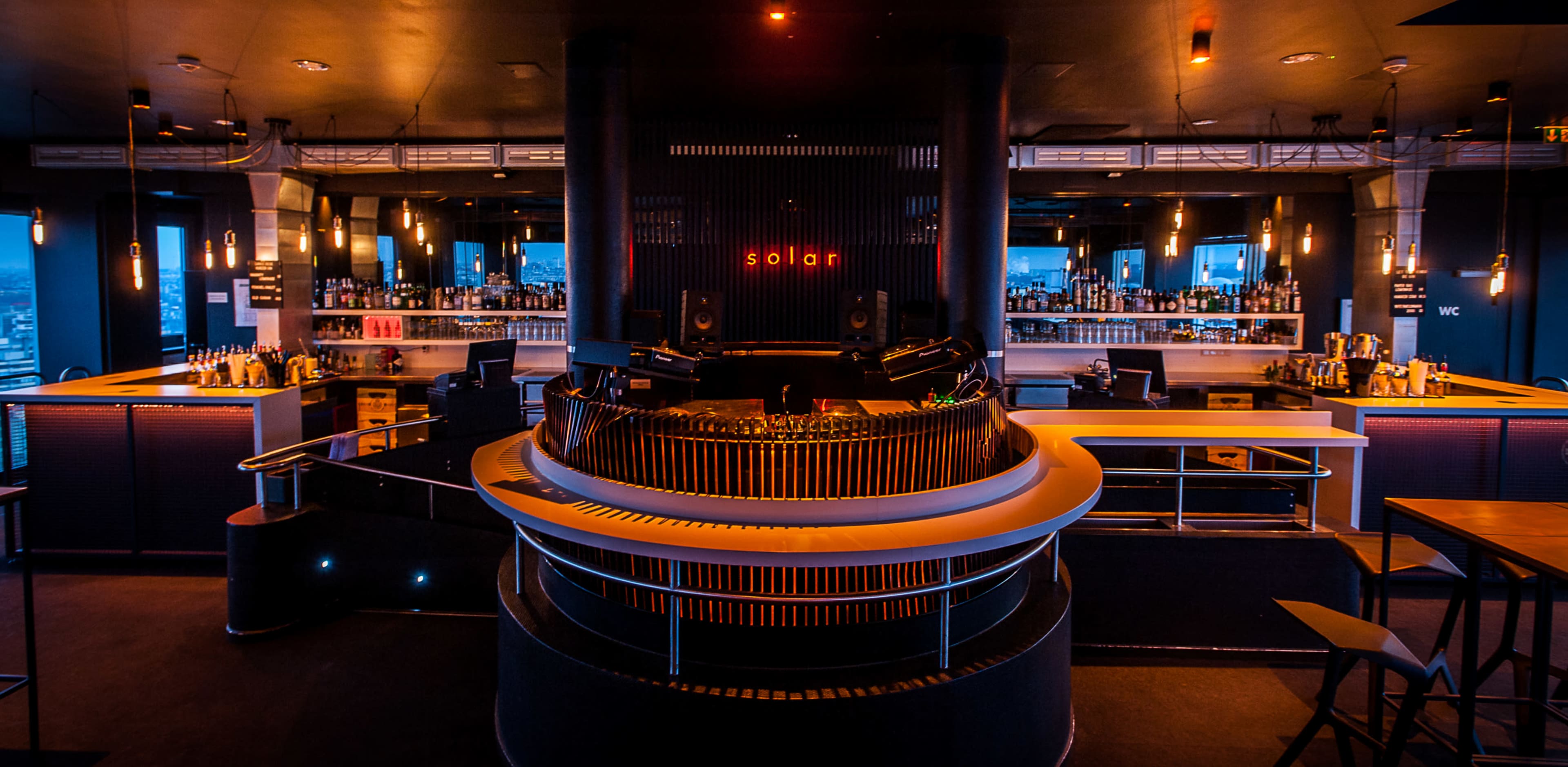 A modern bar interior with a circular bar counter, illuminated by soft lighting and featuring a cityscape view through large windows.