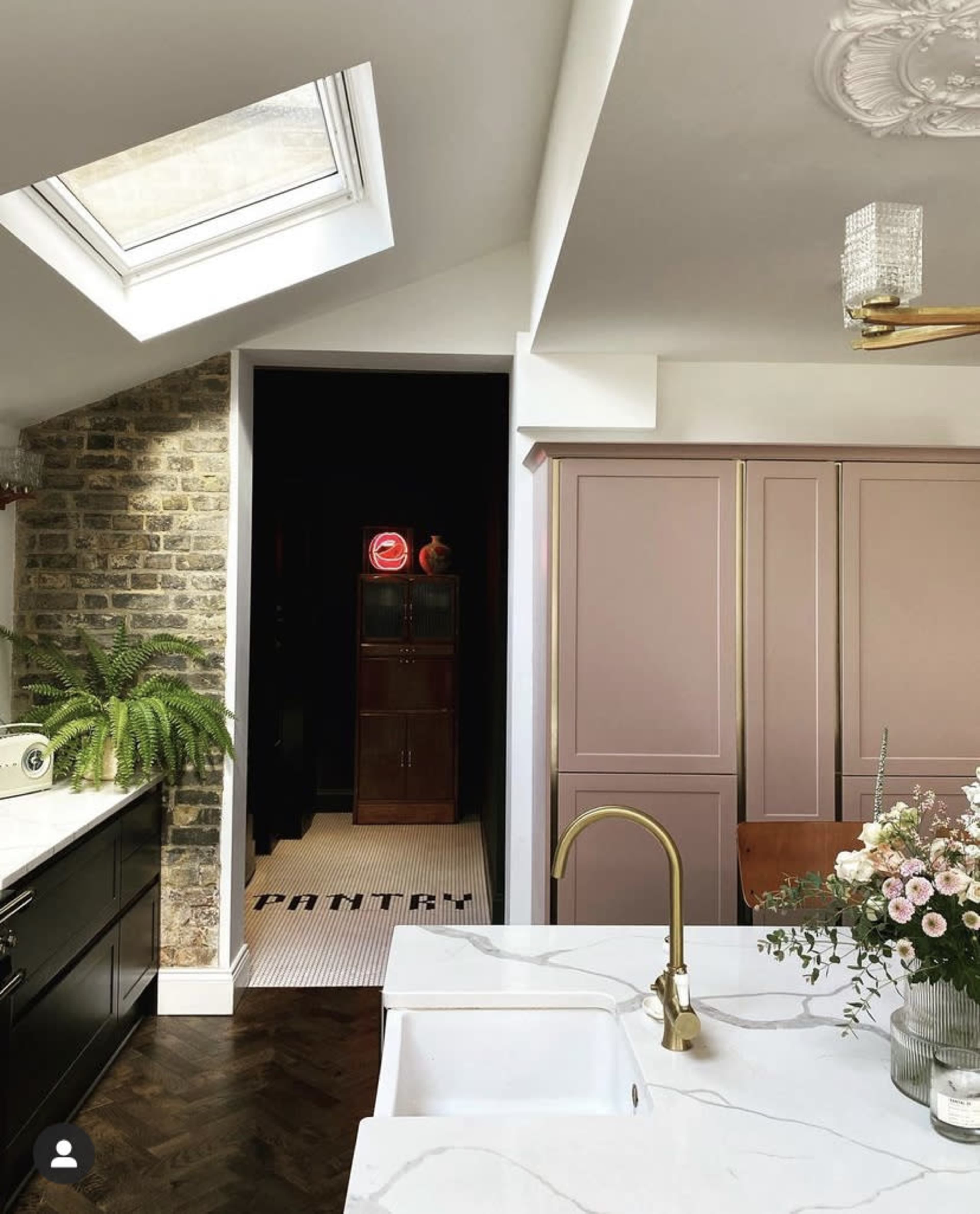A modern kitchen features a skylight, stone walls, and cabinetry in soft hues, with a view leading to a dimly lit hallway.
