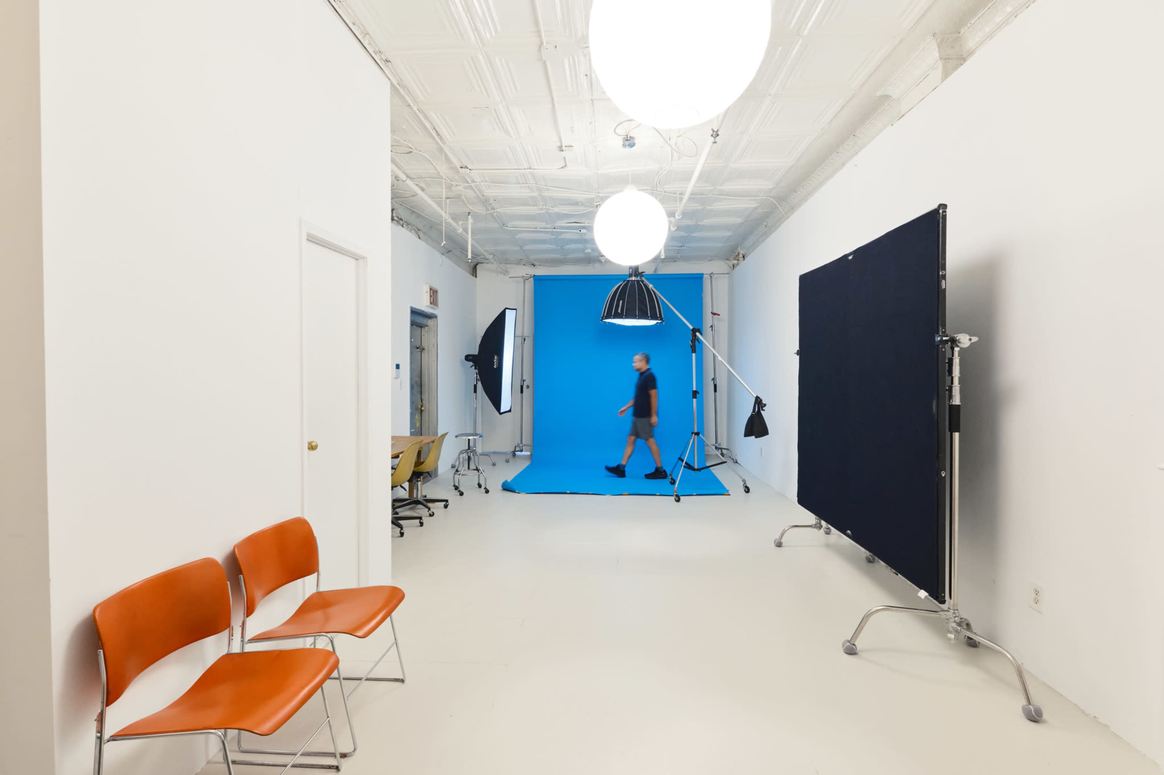 A man walks across a photography studio featuring a blue backdrop, studio lights, and empty chairs.