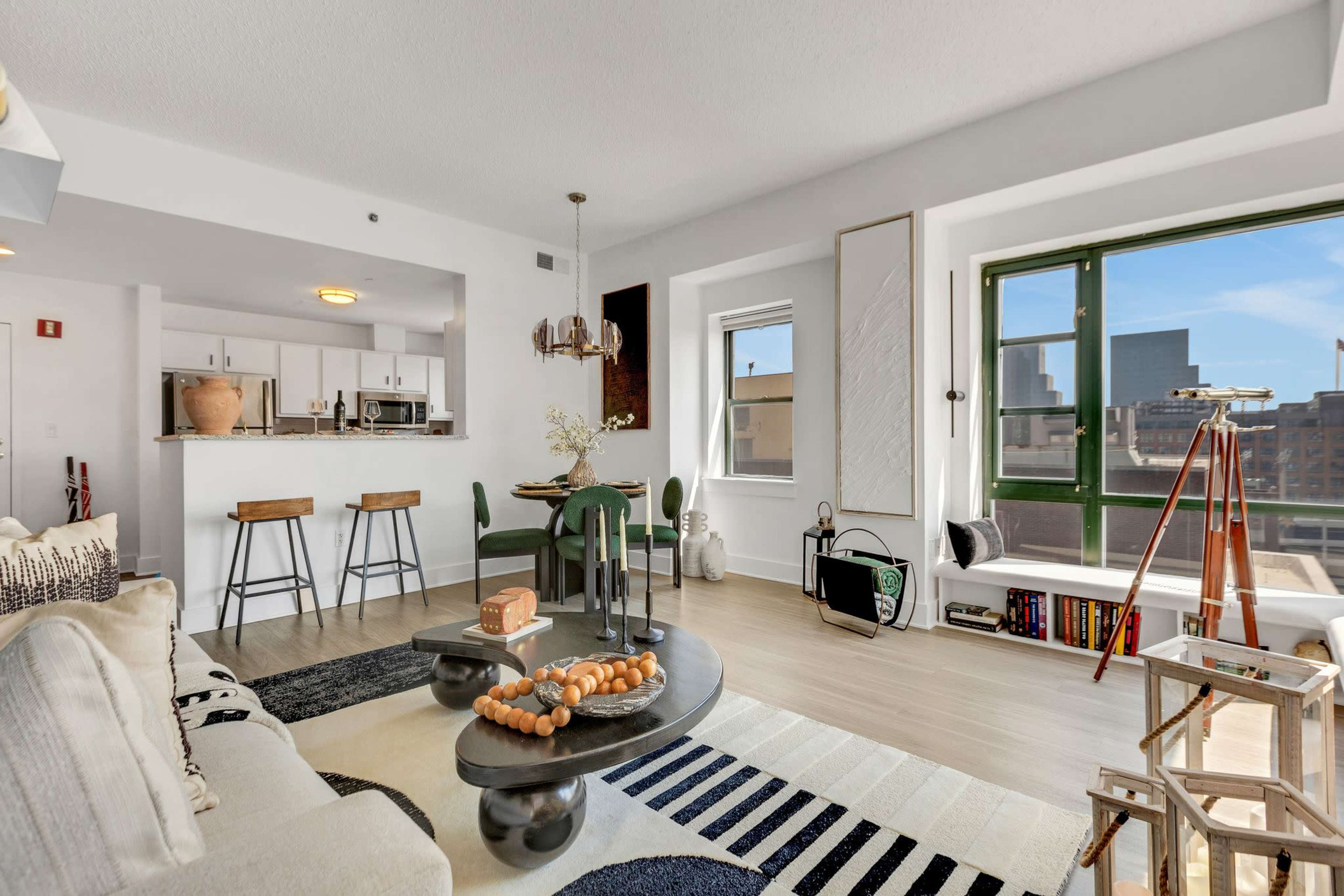 A modern living room area with a round coffee table, green chairs, and a view of city buildings through large windows.