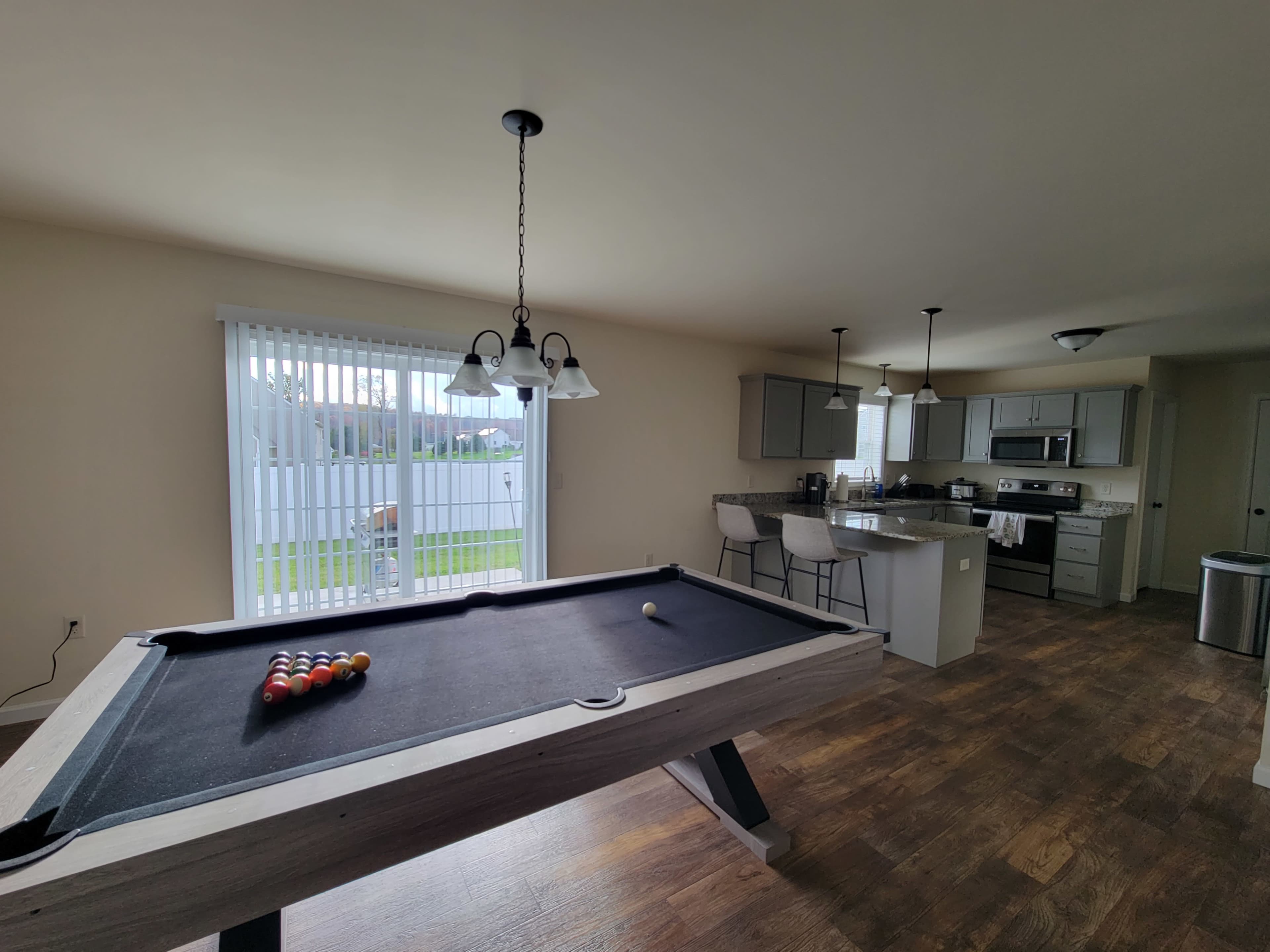 A spacious room featuring a pool table in the foreground and a kitchen with modern appliances in the background, illuminated by natural light from a large window.