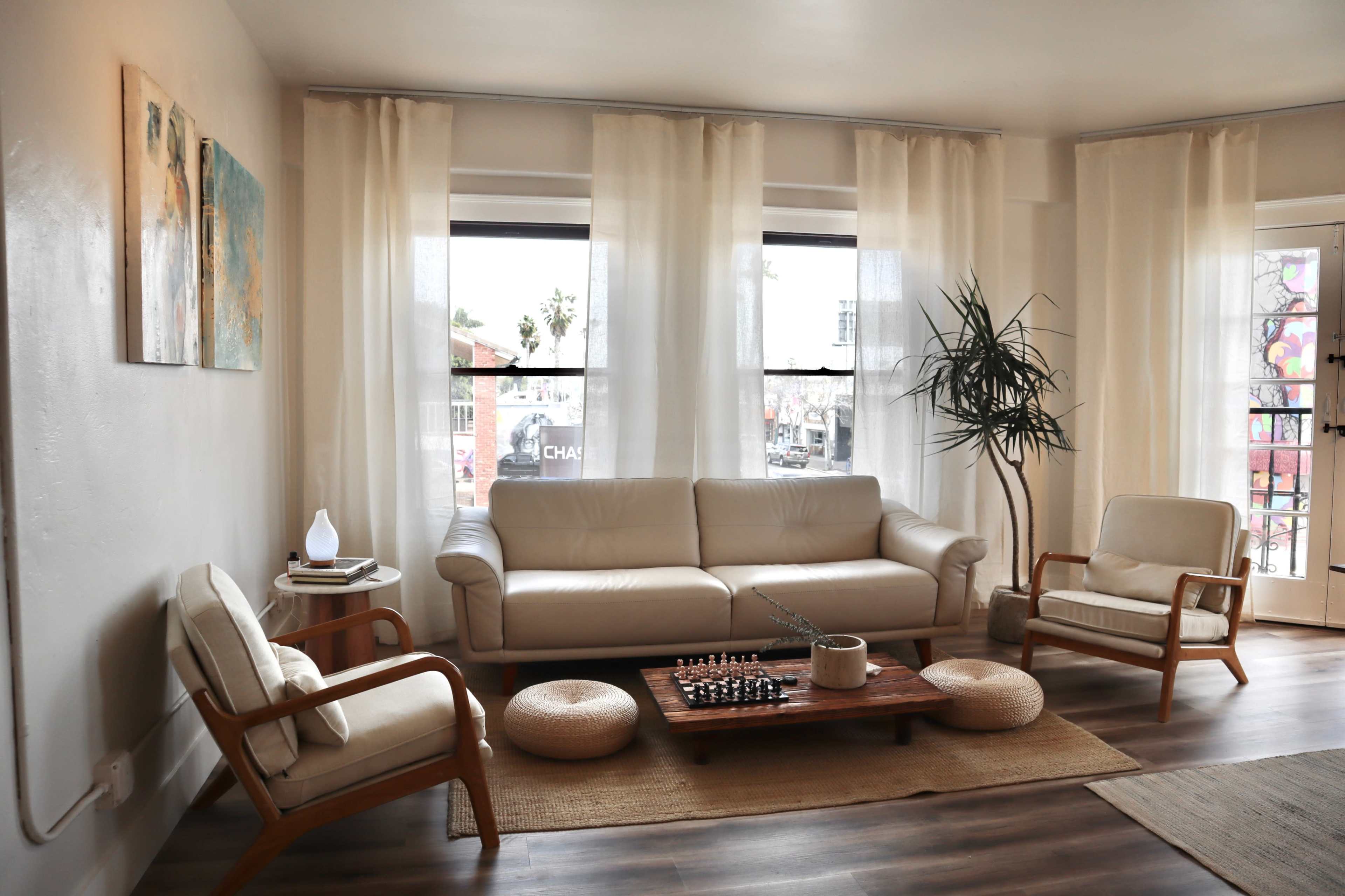 The living room features a light-colored leather sofa, two matching armchairs, a wooden coffee table with a decorative plant, and large windows draped with sheer curtains.