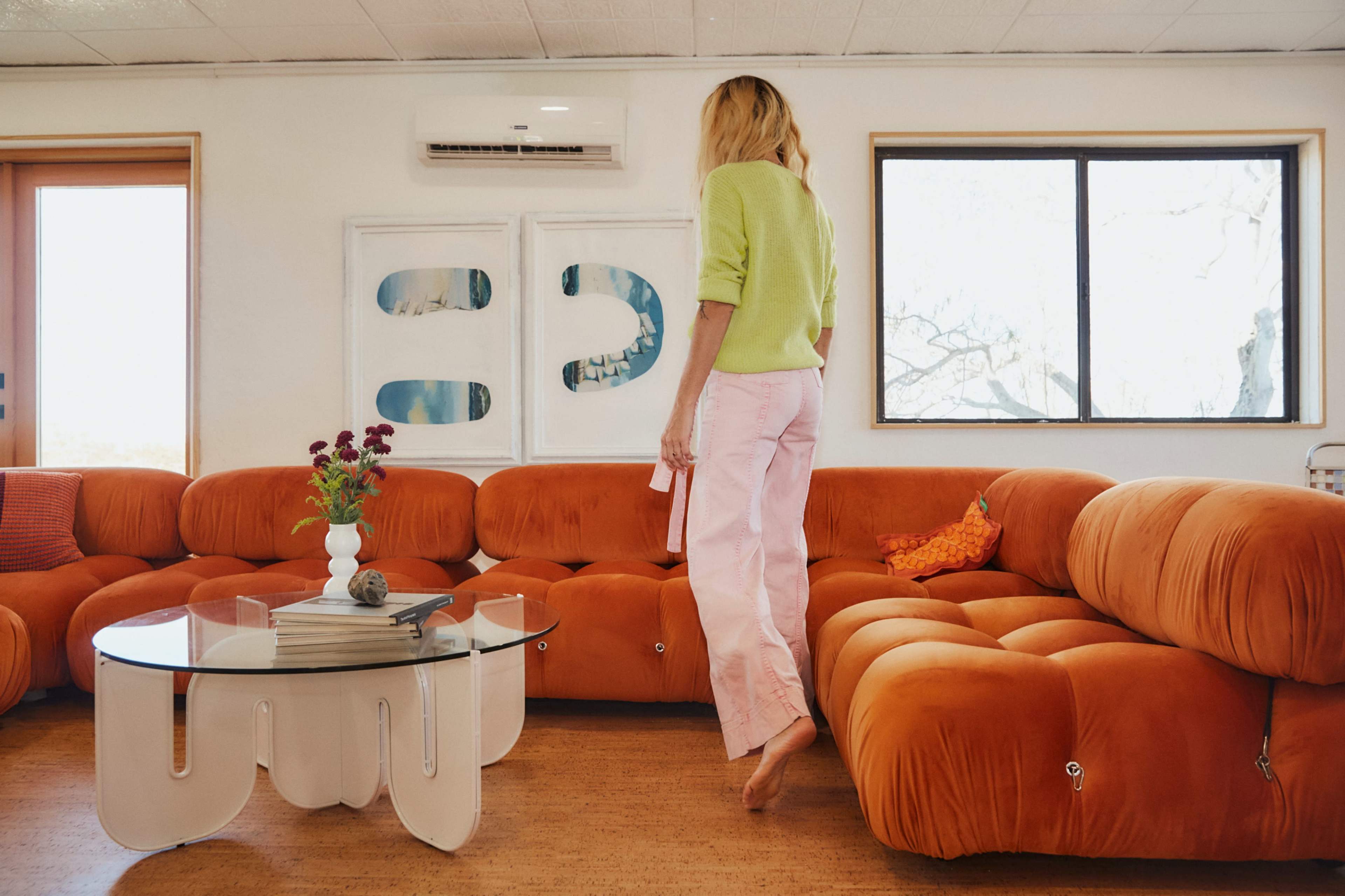 A person walks barefoot on a beige floor beside an orange couch and a glass coffee table in a well-lit room with large windows.