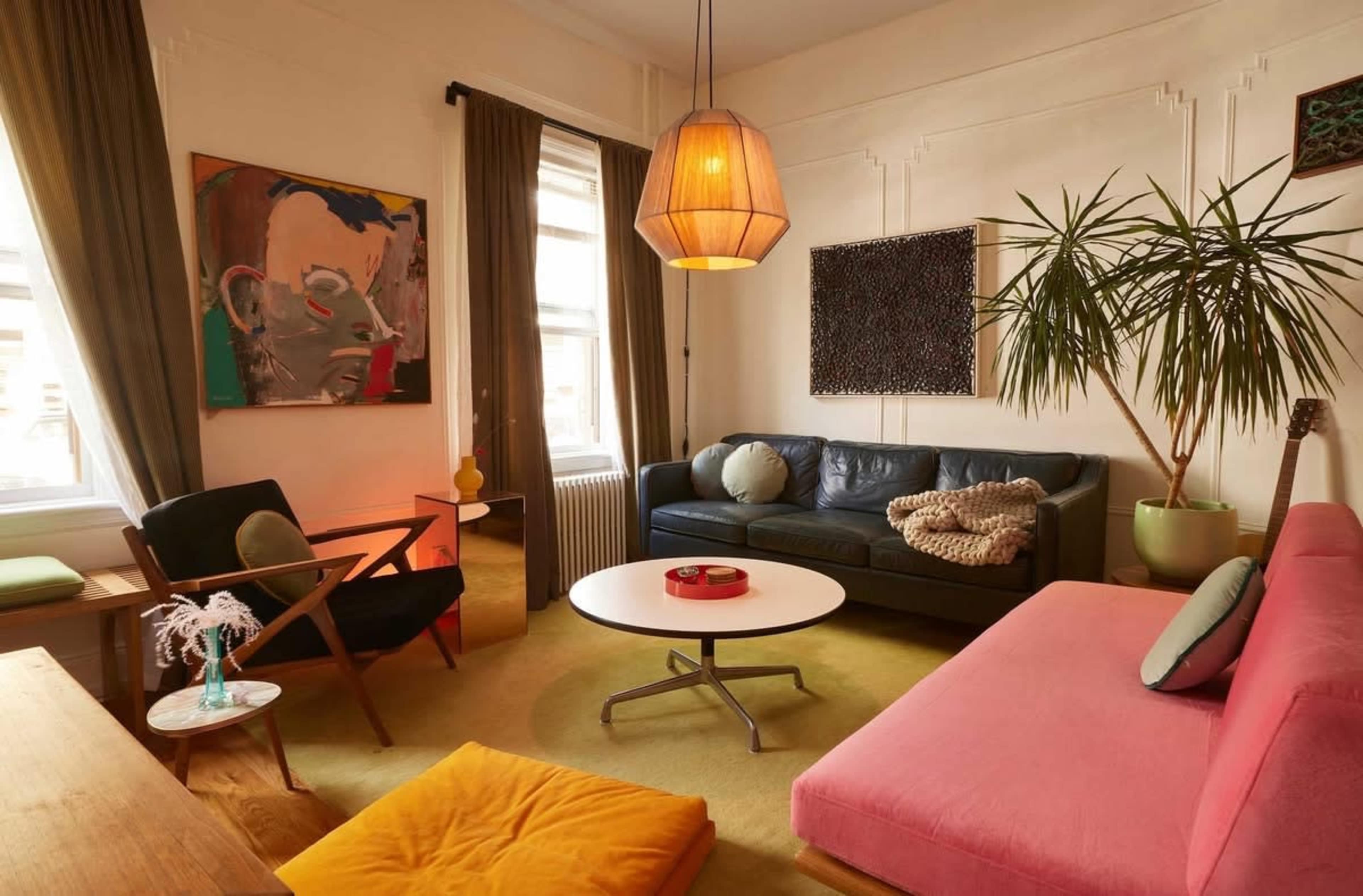 The image shows a living room with a mix of modern furniture, including a black leather sofa, a pink couch, a round coffee table, and decorative plants, along with colorful artwork on the walls.