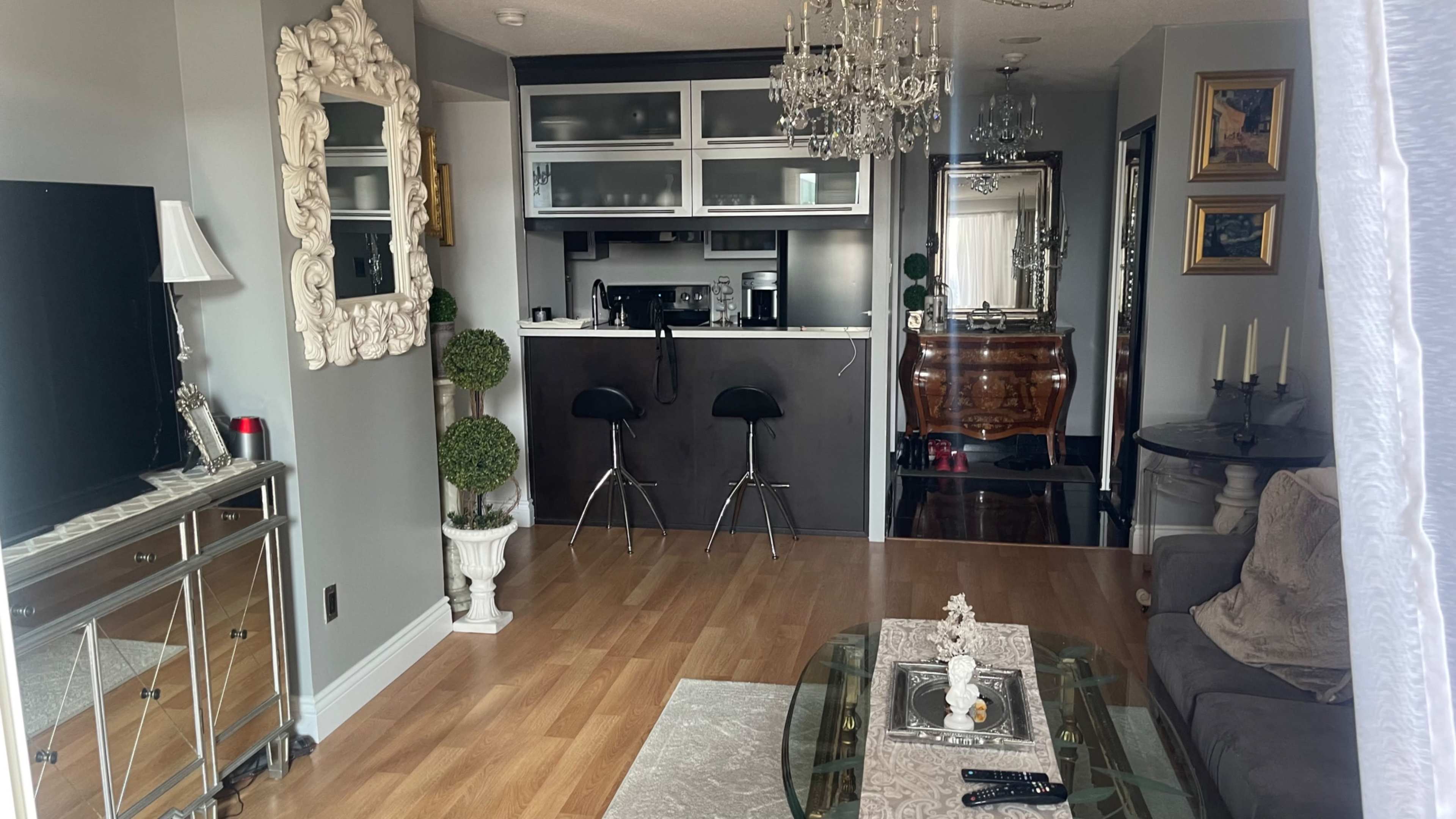The image shows a modern living area with a kitchen bar, a glass coffee table, and decorative elements like plants and a chandelier.
