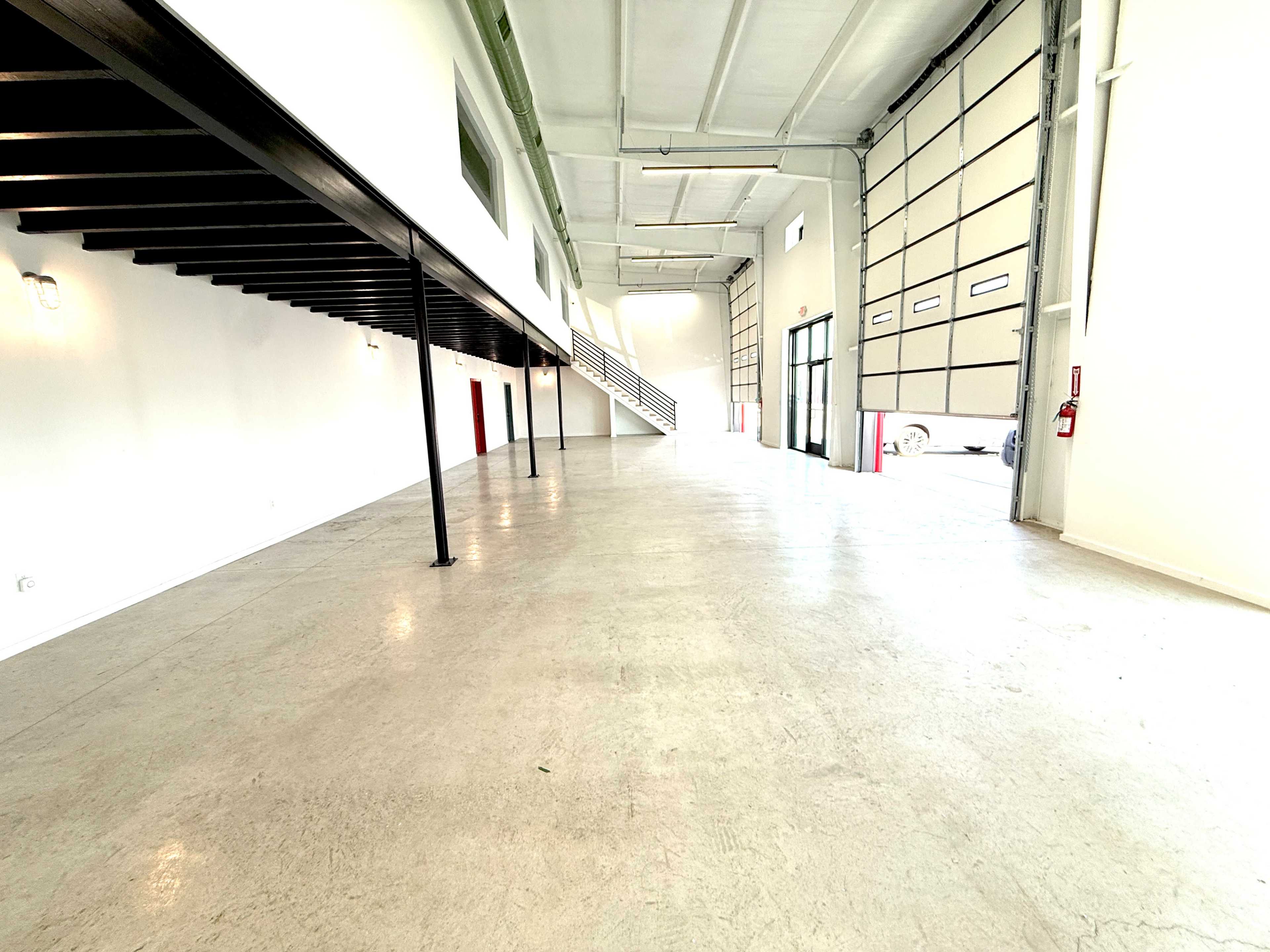 The image shows an empty industrial space with concrete flooring, metal support beams, and large garage doors.