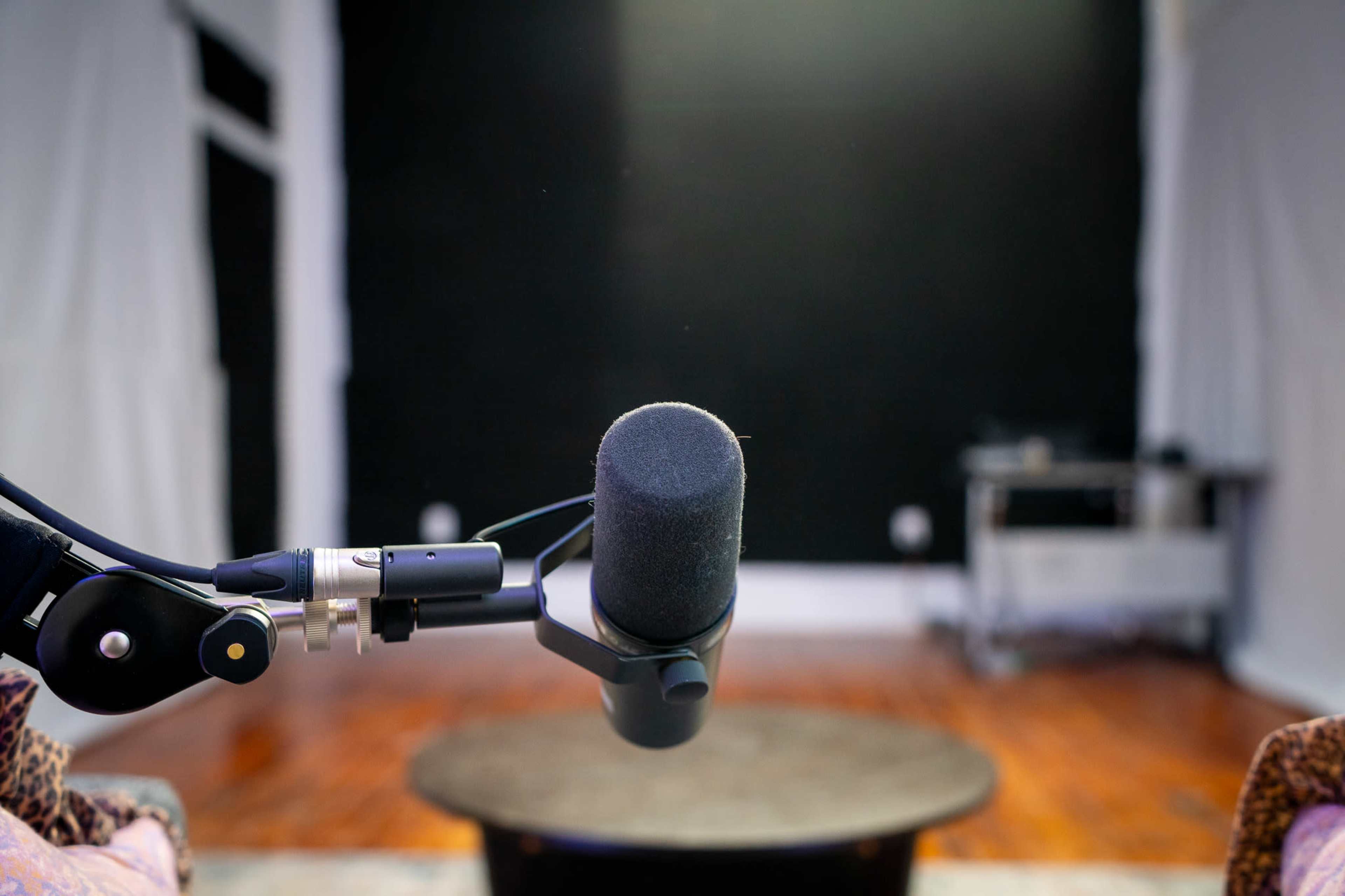 A microphone is positioned in the foreground, facing a round table in a sparsely furnished room with a dark wall.