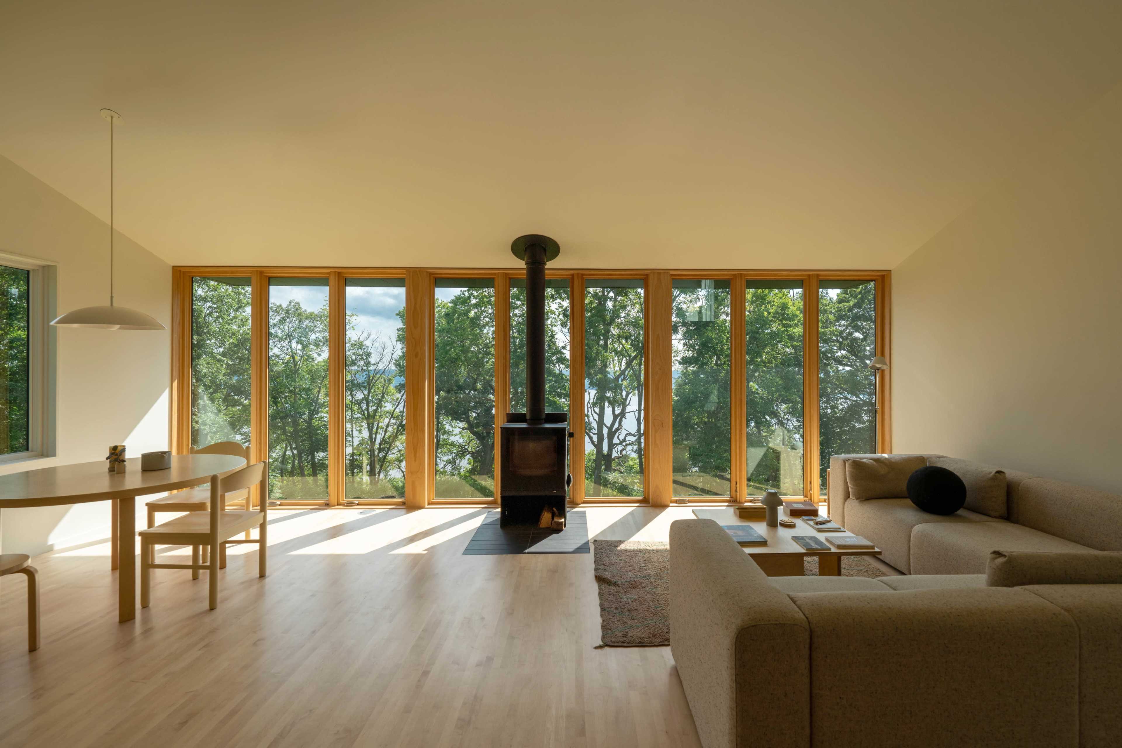 The image shows a modern living room with a large window wall, allowing natural light to illuminate the space, and features a seating area with a sofa, a round dining table, and a wood stove.