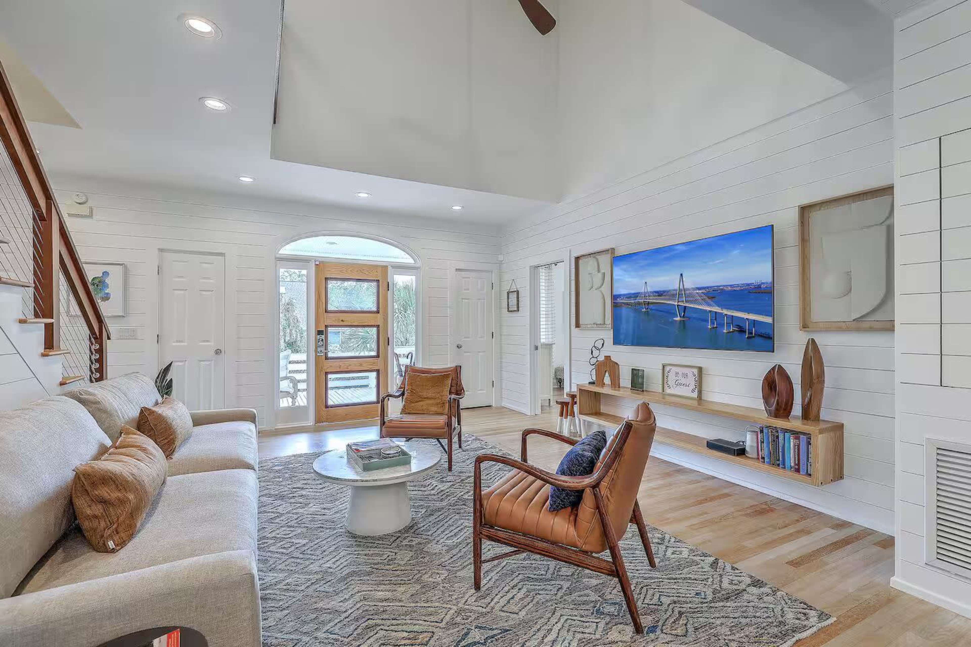 The image shows a modern living room with a light-colored sofa, wooden chairs, and a large flat-screen television mounted on the wall, alongside a coffee table and wooden shelves displaying decorative items.