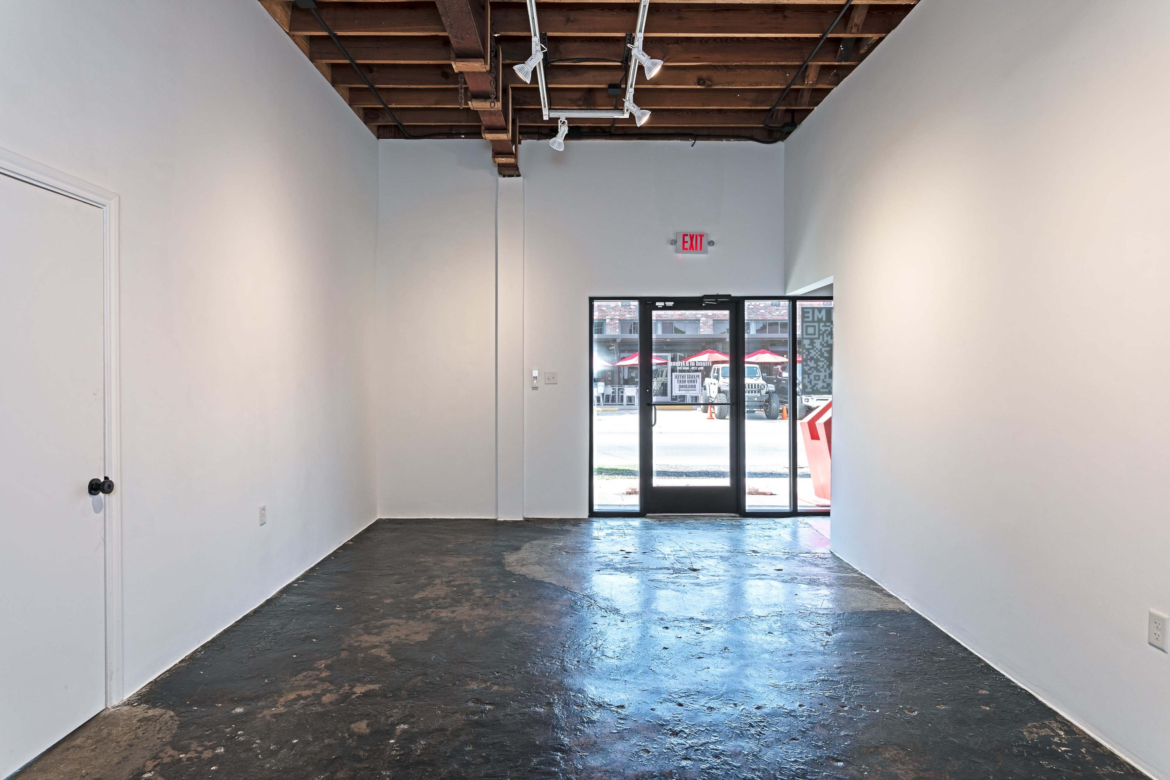A minimalist gallery space with white walls, a concrete floor, and a glass door leading outside.