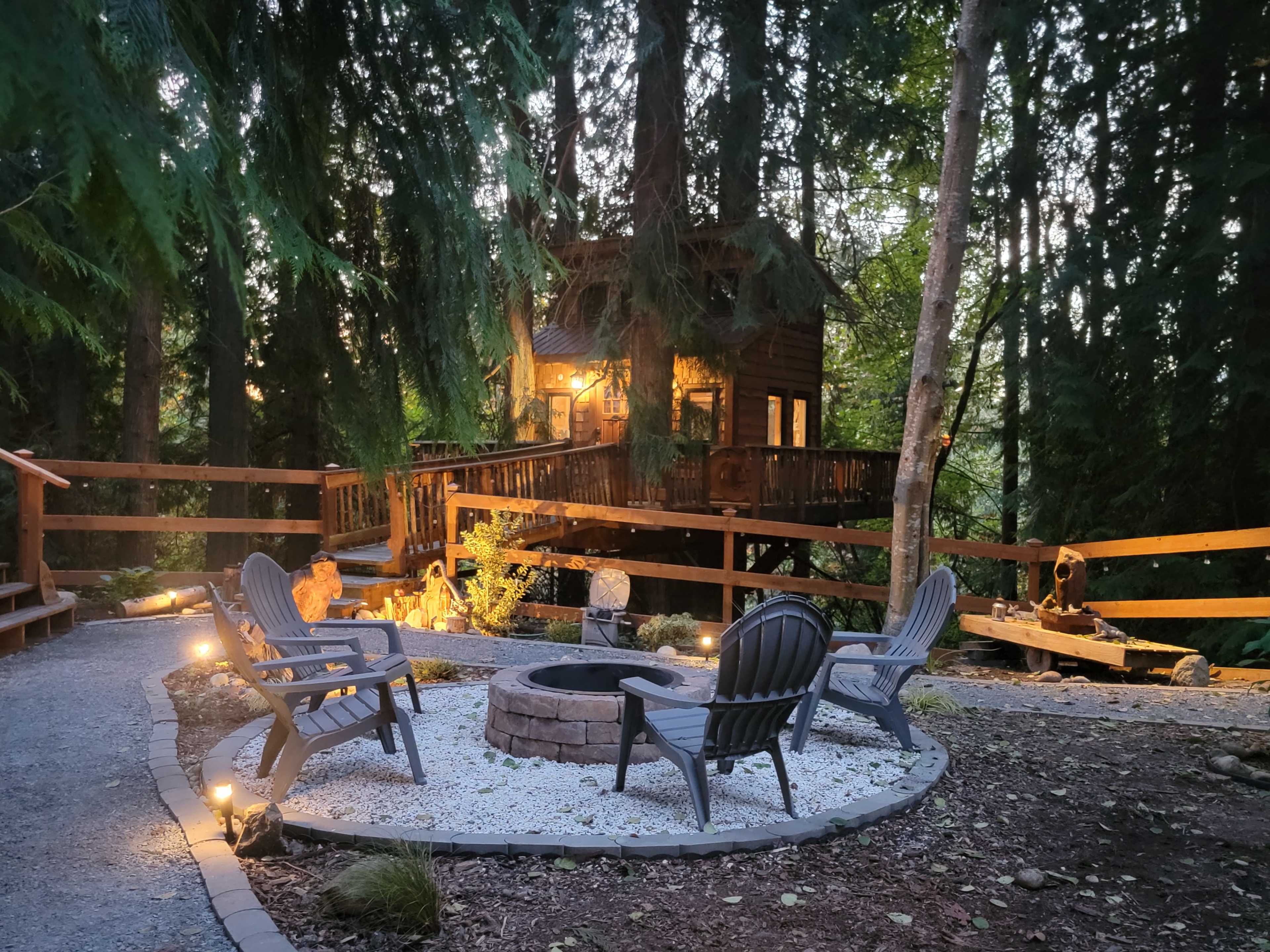 The scene features a fire pit surrounded by four chairs, with a treehouse visible in the background among tall trees.