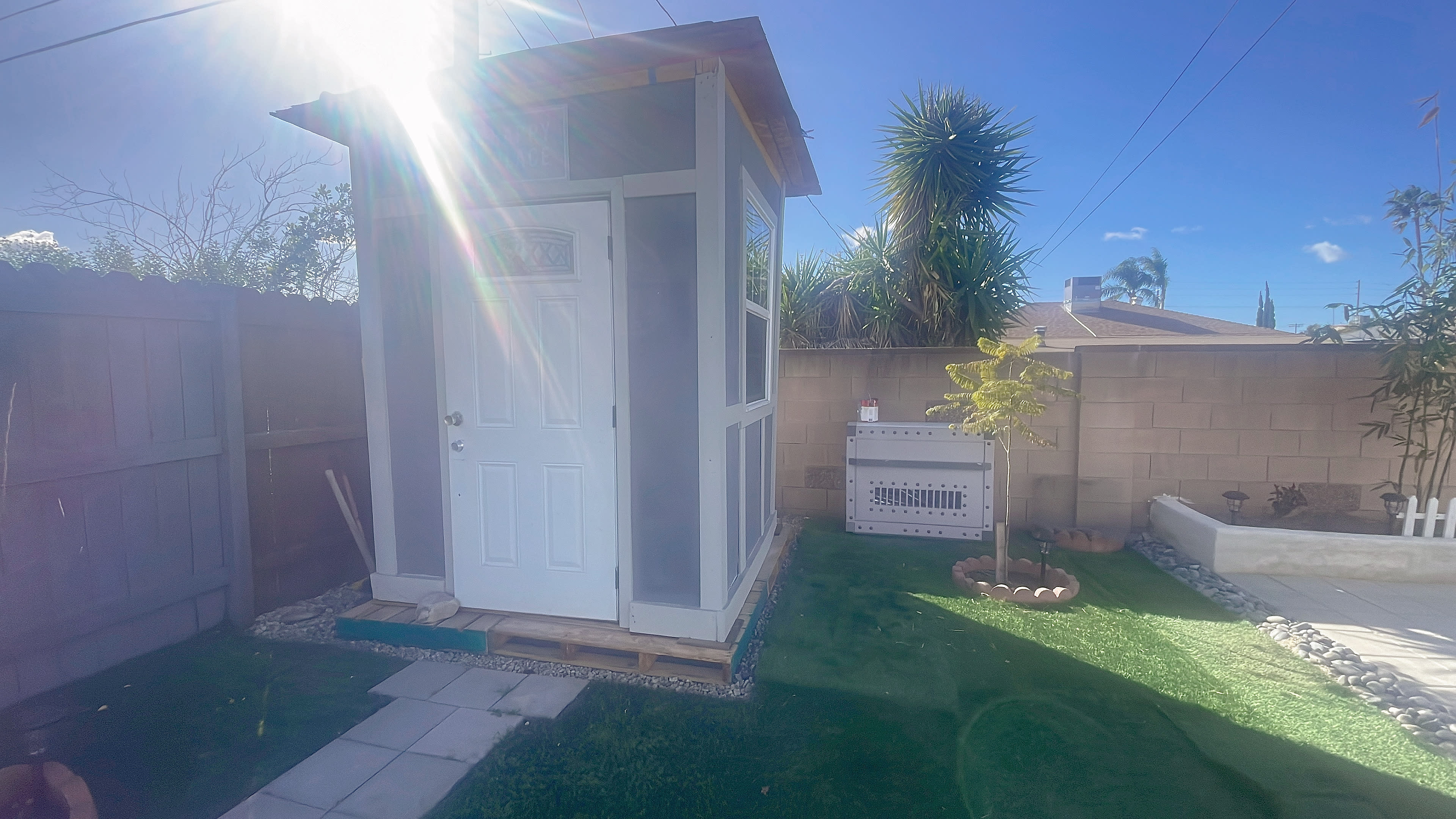 A small white shed with a door and windows stands beside an outdoor air conditioning unit in a grassy yard.