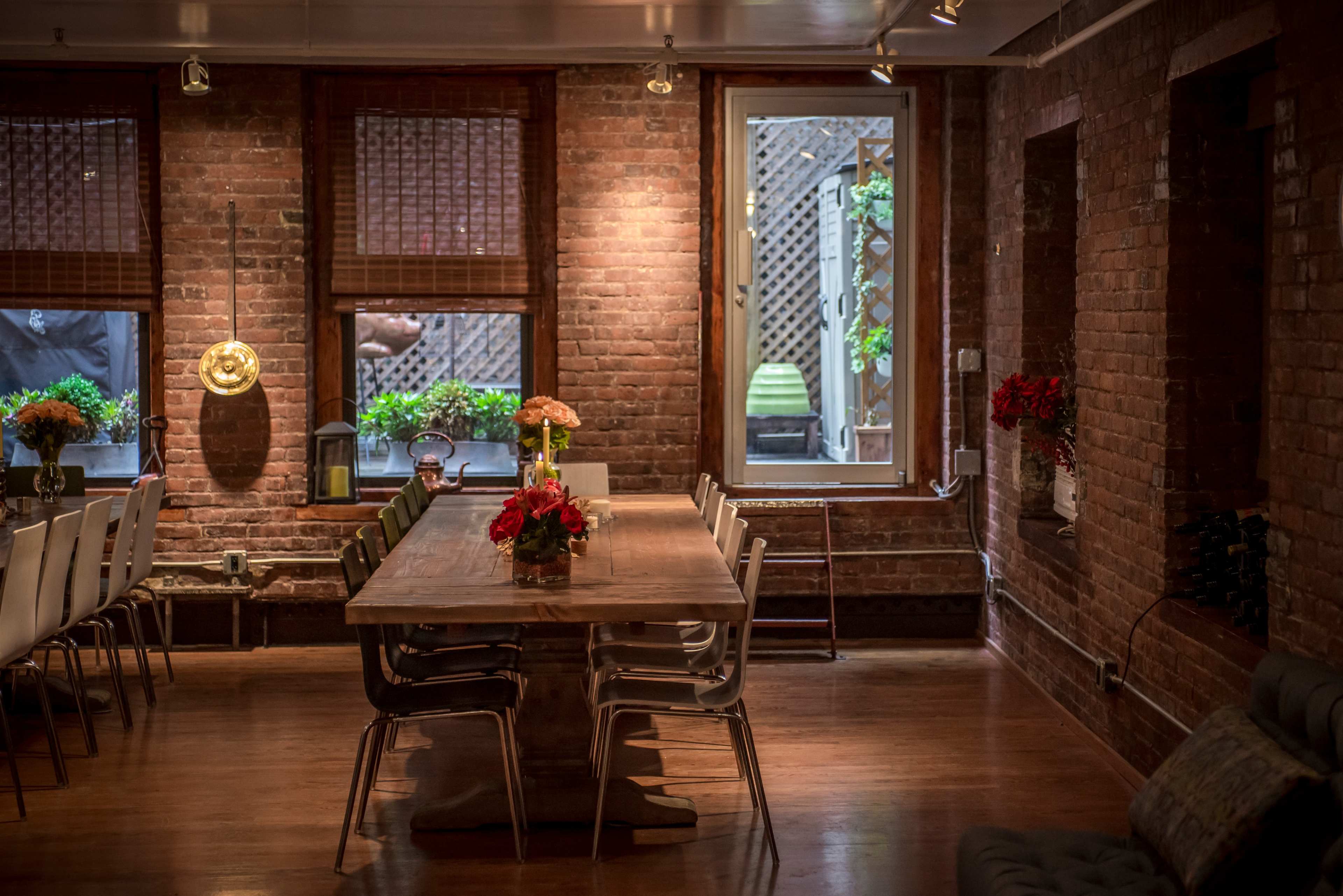 A dining area with a large wooden table surrounded by chairs, set against a backdrop of exposed brick walls and windows overlooking a small garden.