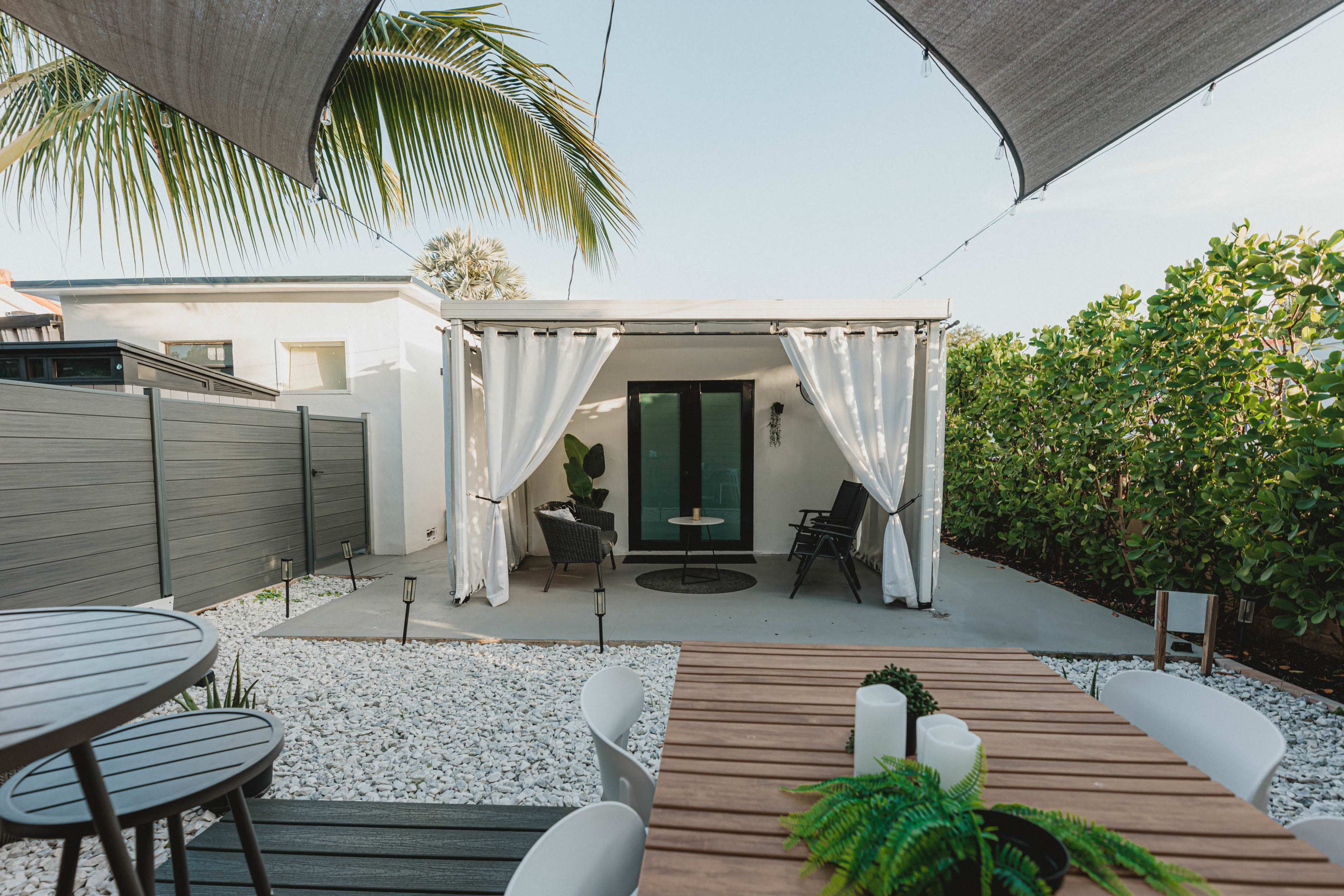 The image shows a modern outdoor patio area with a seating arrangement, a small table, and a structure partially surrounded by greenery and gravel.