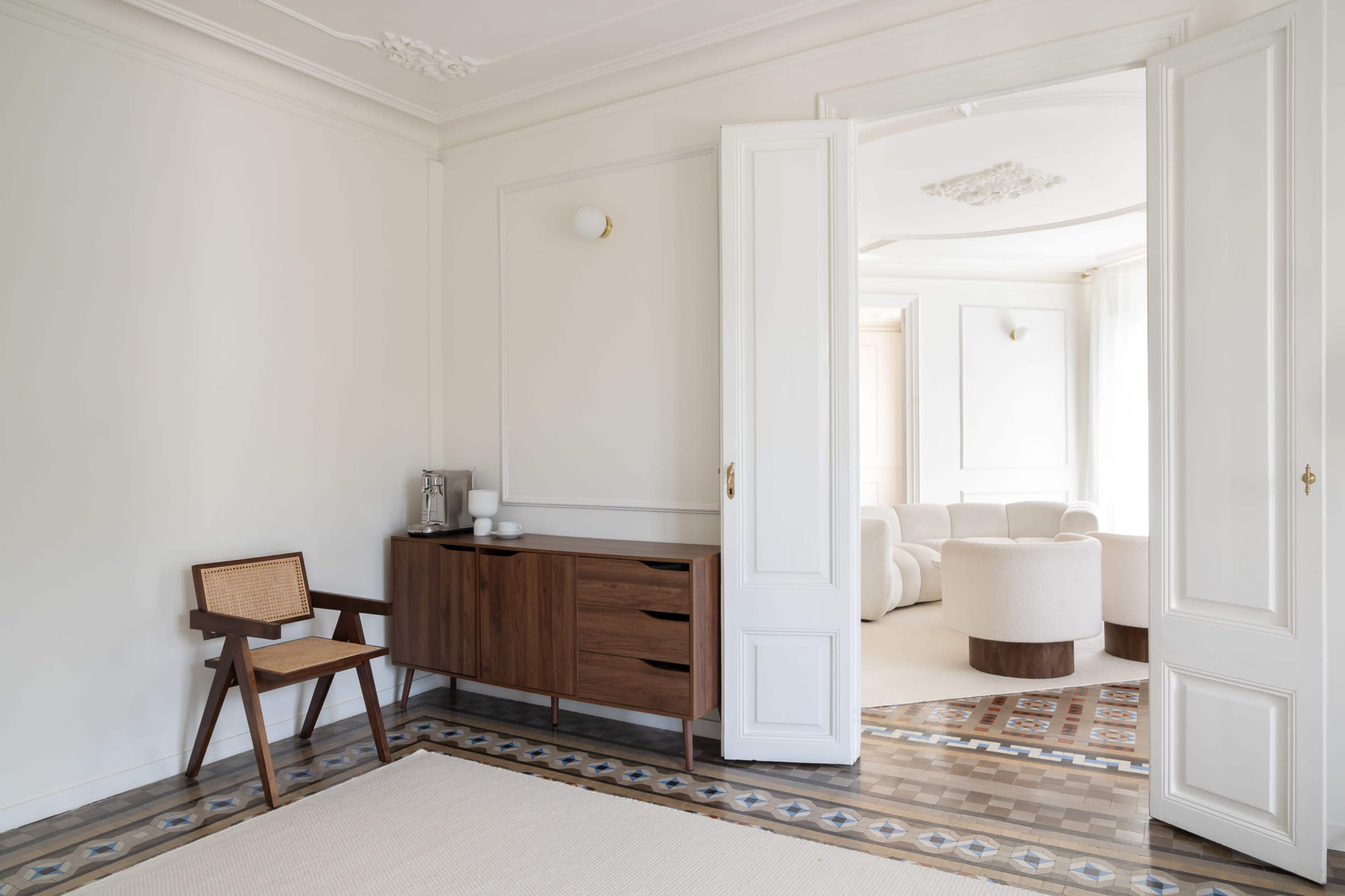 A spacious room features a wooden sideboard with a chair, leading to a bright area with circular furniture.