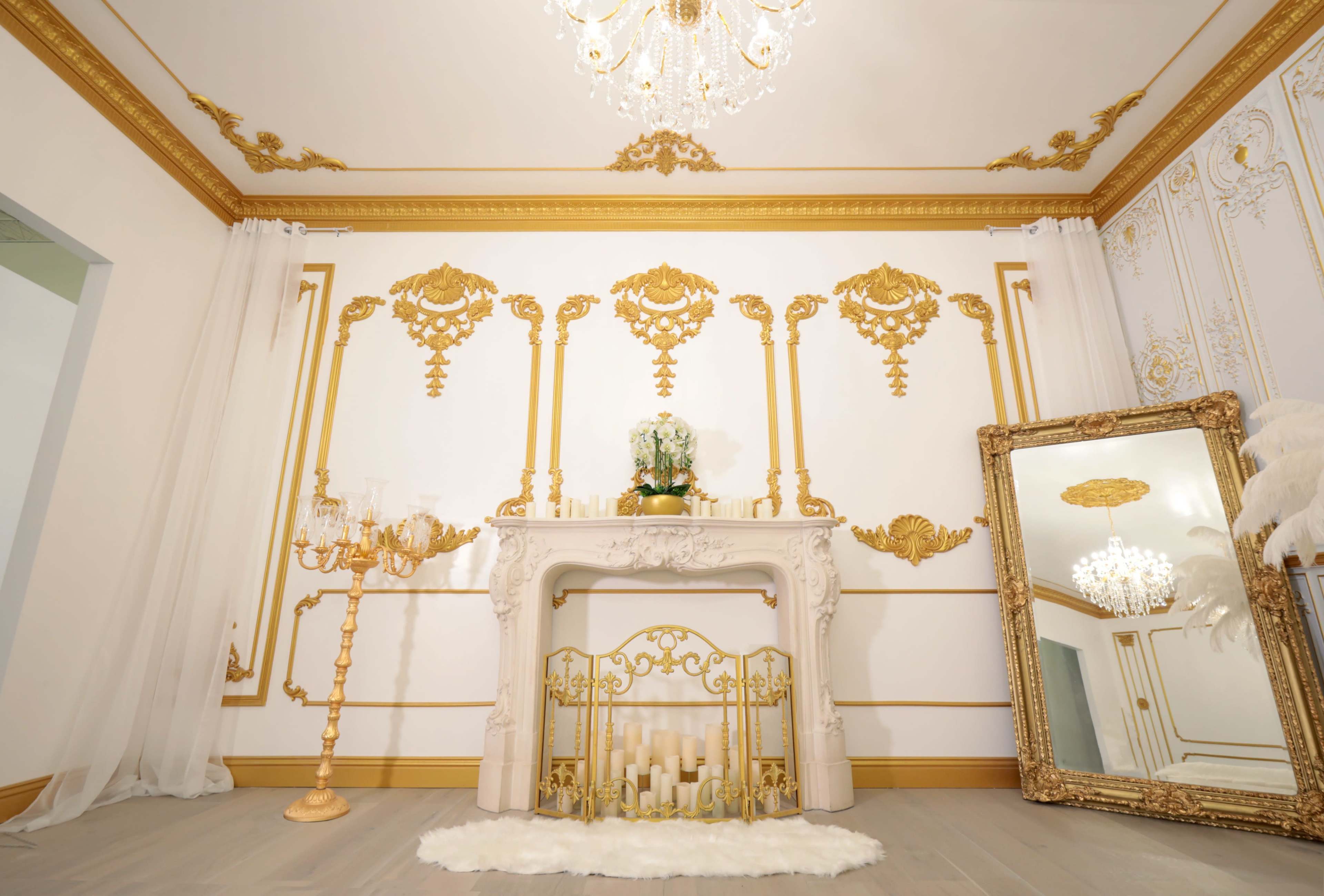 An elegantly decorated room featuring a white and gold ornamental fireplace, a large ornate mirror, candles, and a chandelier.