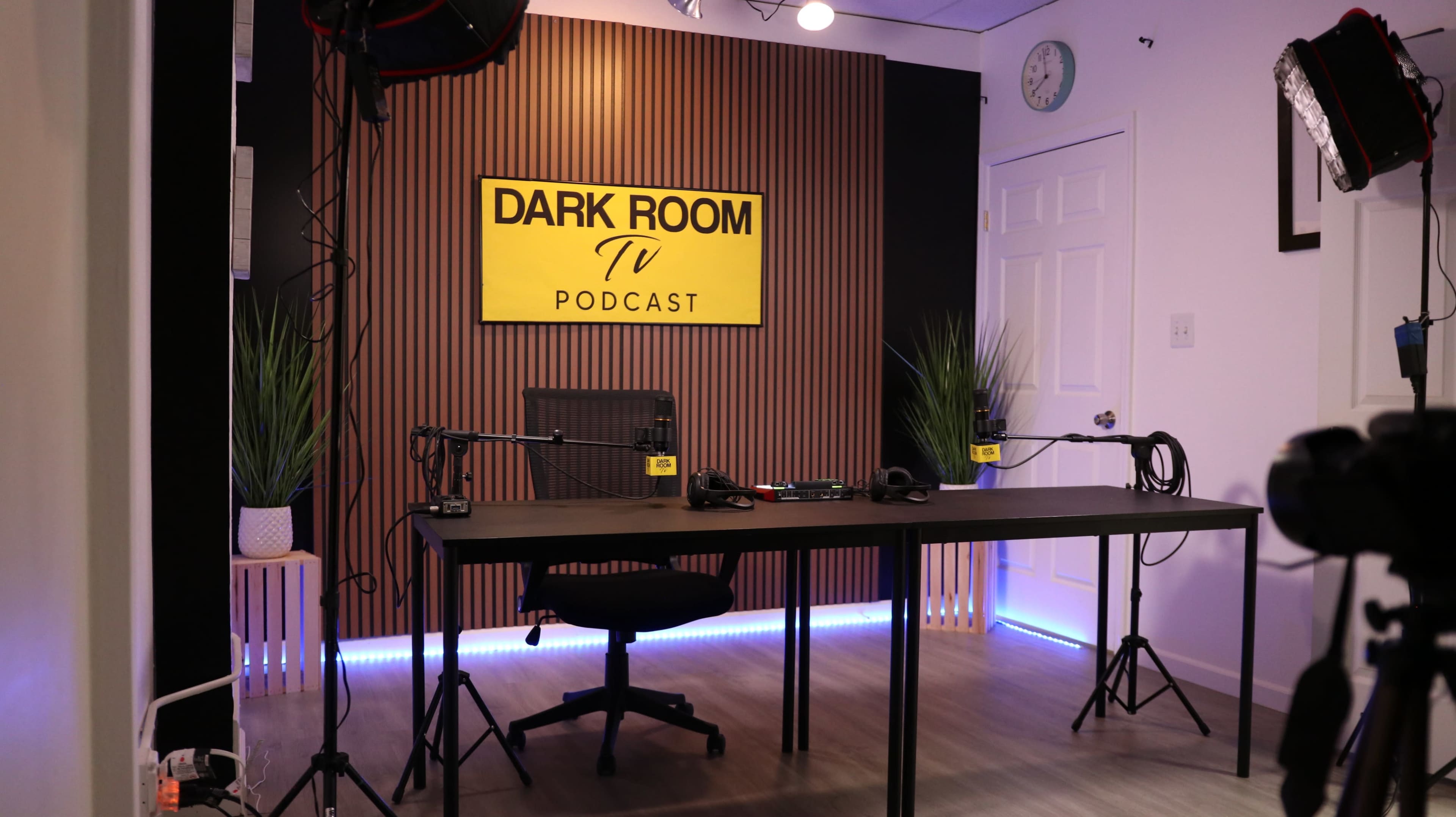 The image shows a well-lit podcast studio featuring a large yellow sign reading "DARK ROOM TV PODCAST," with a central table, a rolling chair, and microphones set up on stands.