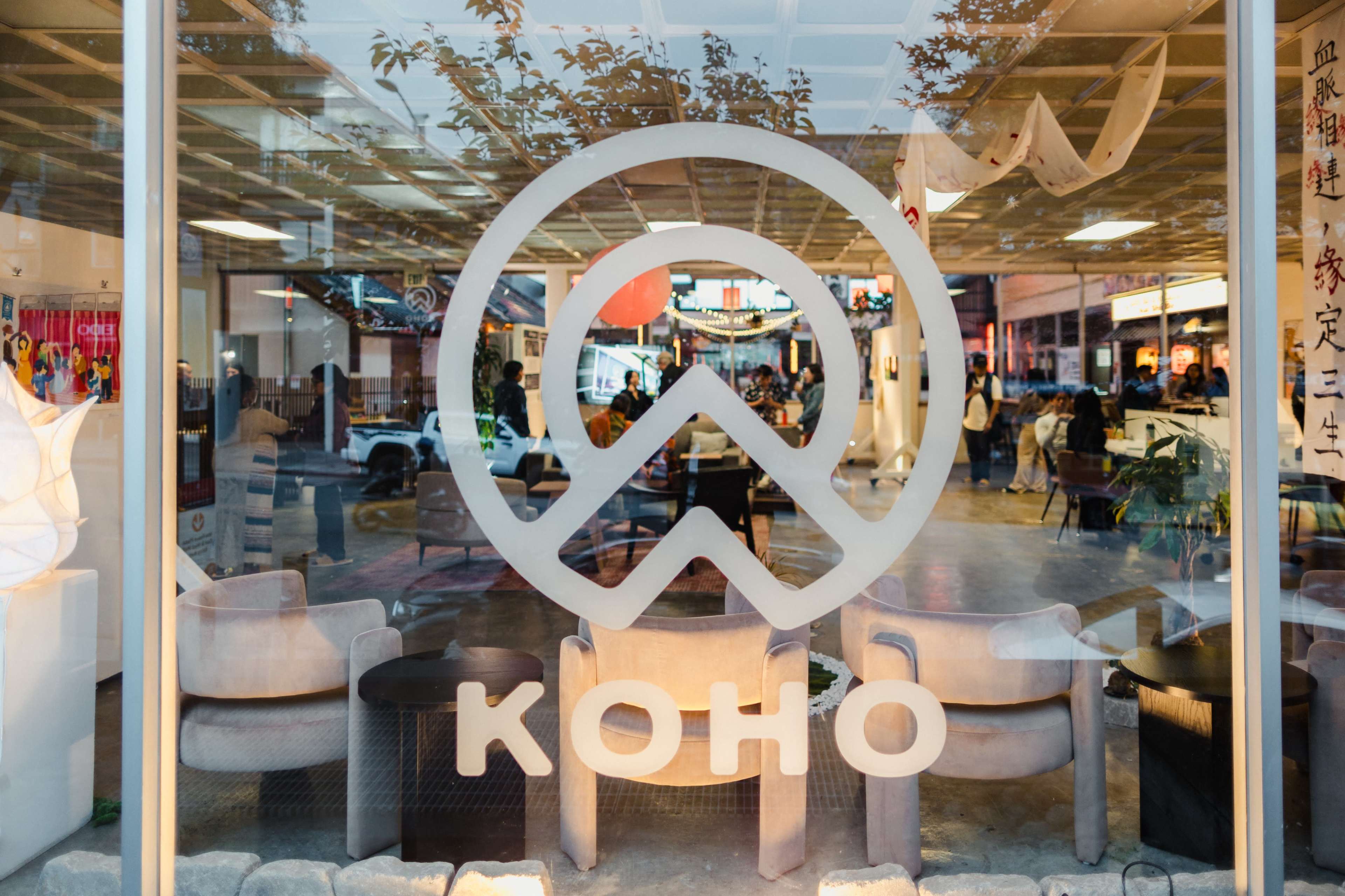 A large, stylized logo of "KOHO" is displayed on a glass window, reflecting the interior of a bustling space with people and decor in the background.