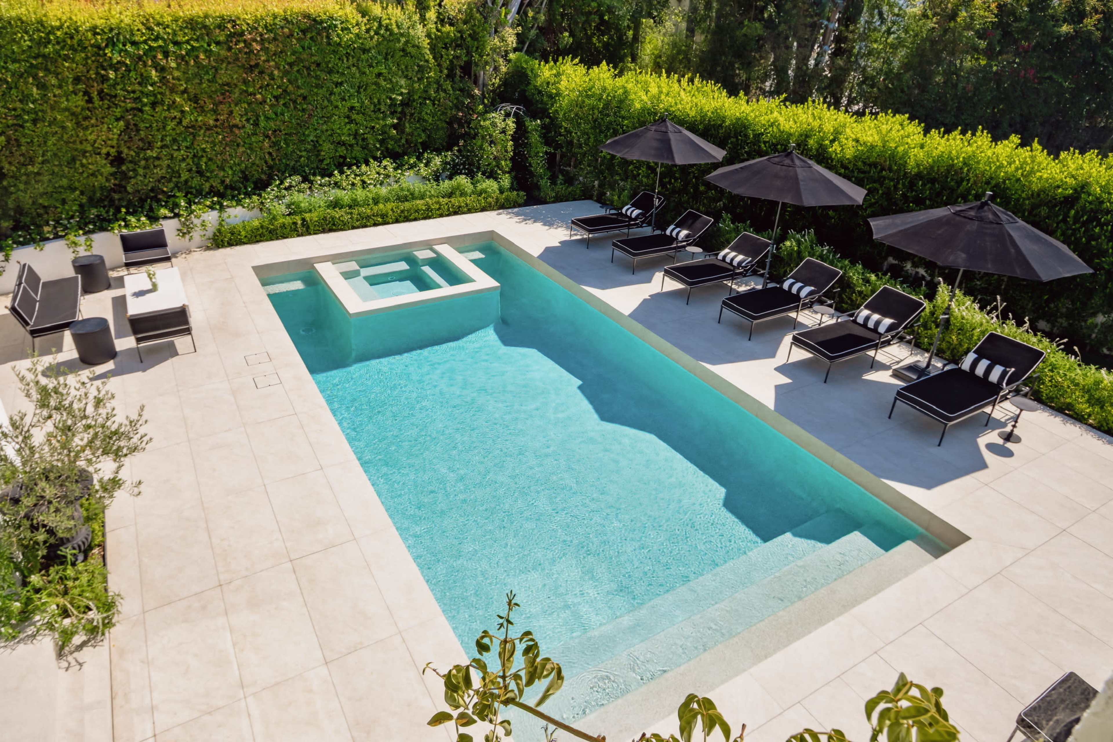 The image shows a modern swimming pool surrounded by lounge chairs and umbrellas in a landscaped outdoor setting.