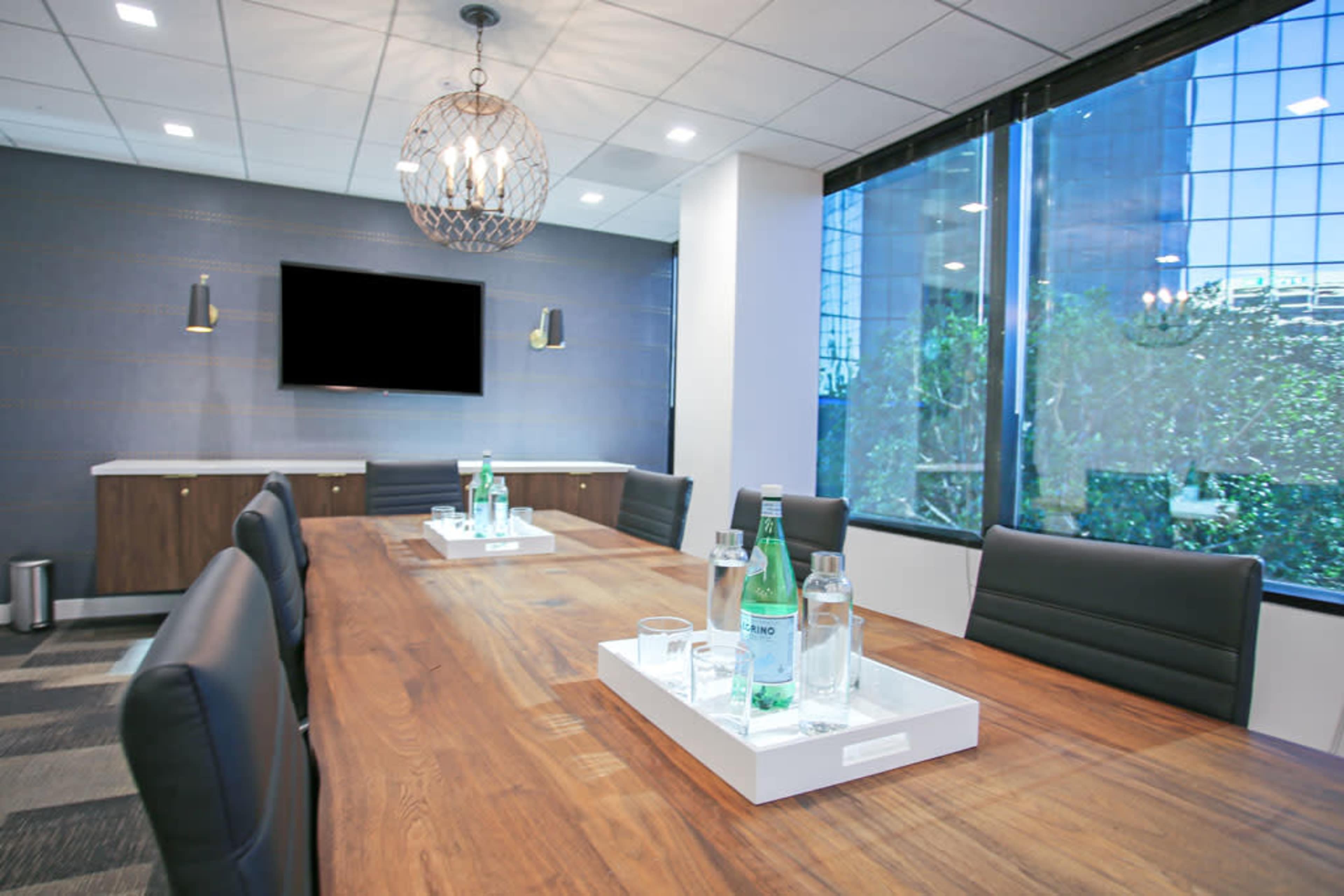 A modern conference room features a large wooden table with water bottles and glasses, surrounded by black leather chairs and a large window displaying a view of greenery outside.