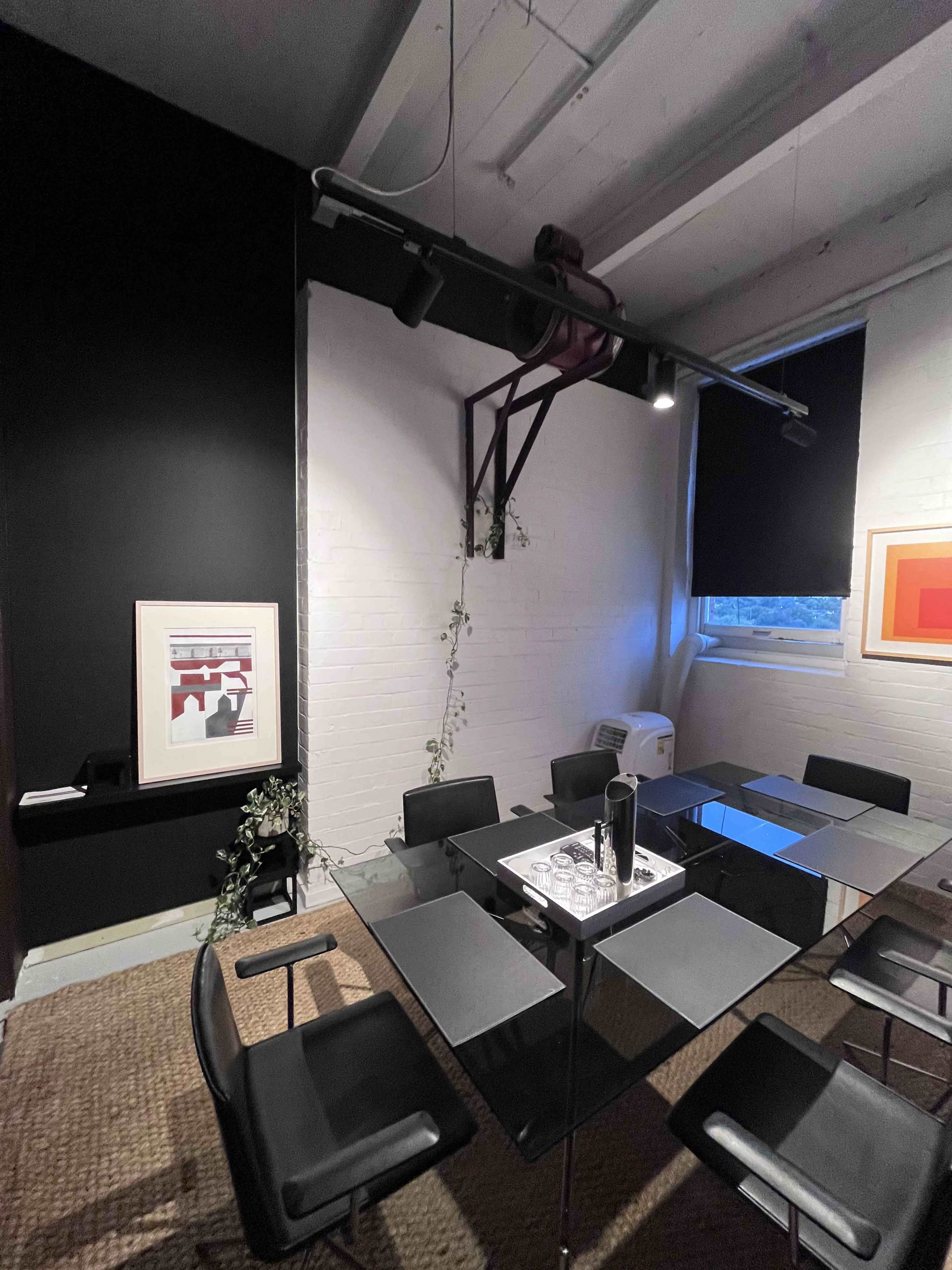 The image shows a modern conference room with a glass table surrounded by black chairs, a white brick wall adorned with artwork, and a plant along the side.
