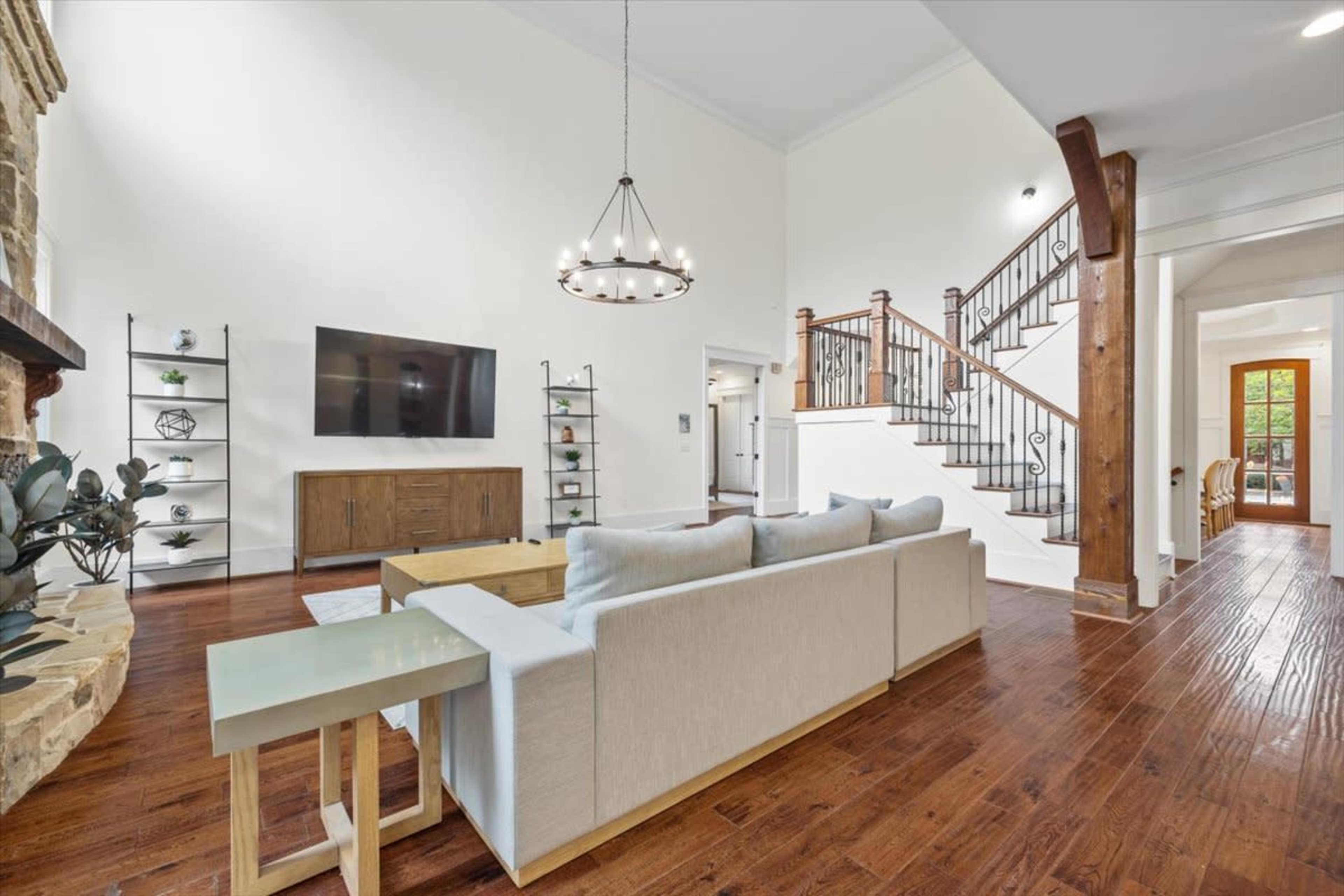 A modern living room features a light-colored sofa facing a wall-mounted television, with a wooden cabinet, a staircase, and large windows in the background.