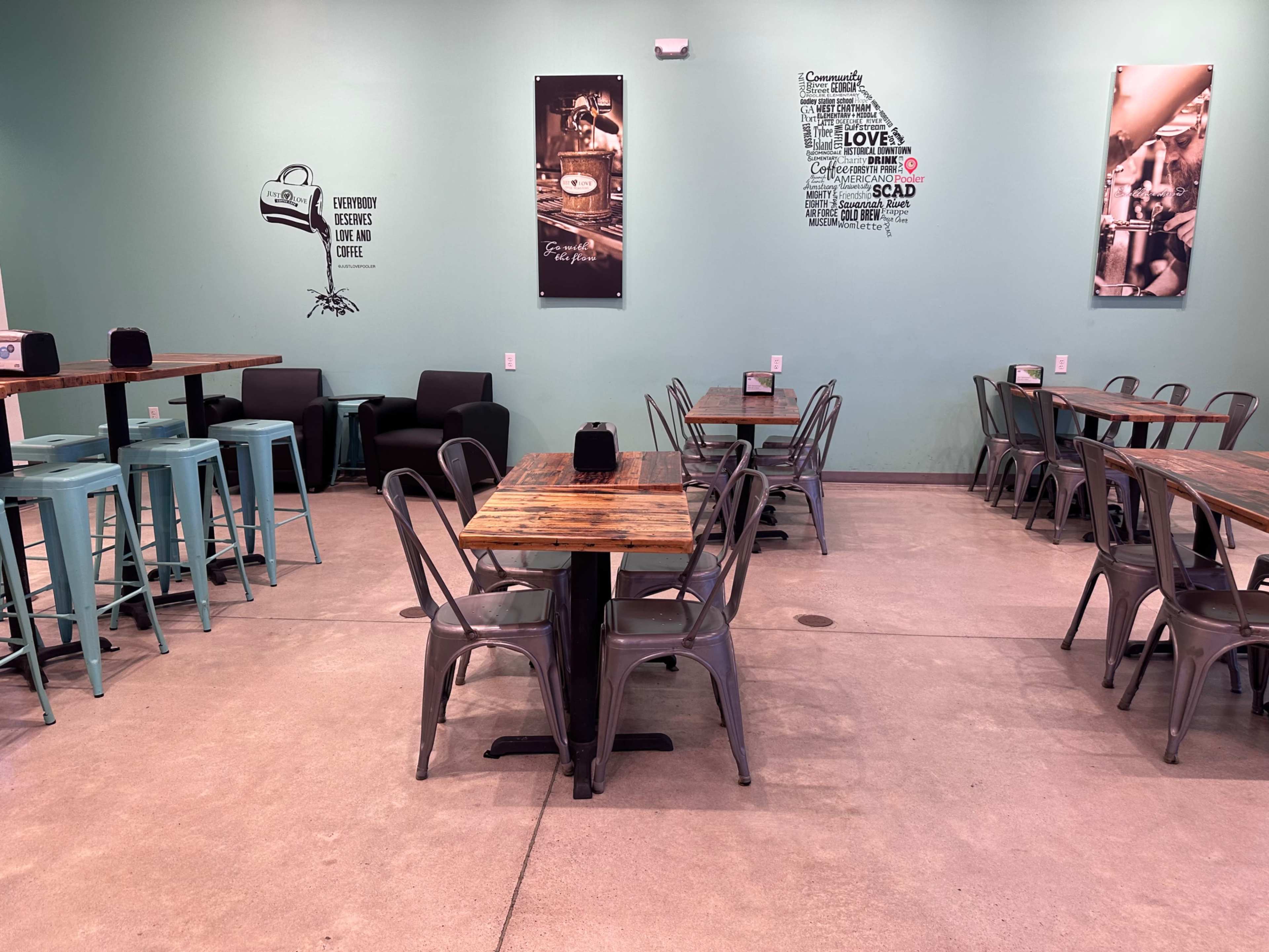 A coffee shop interior with metal tables and chairs arranged in a spacious area, featuring wall decorations and a green-painted wall.