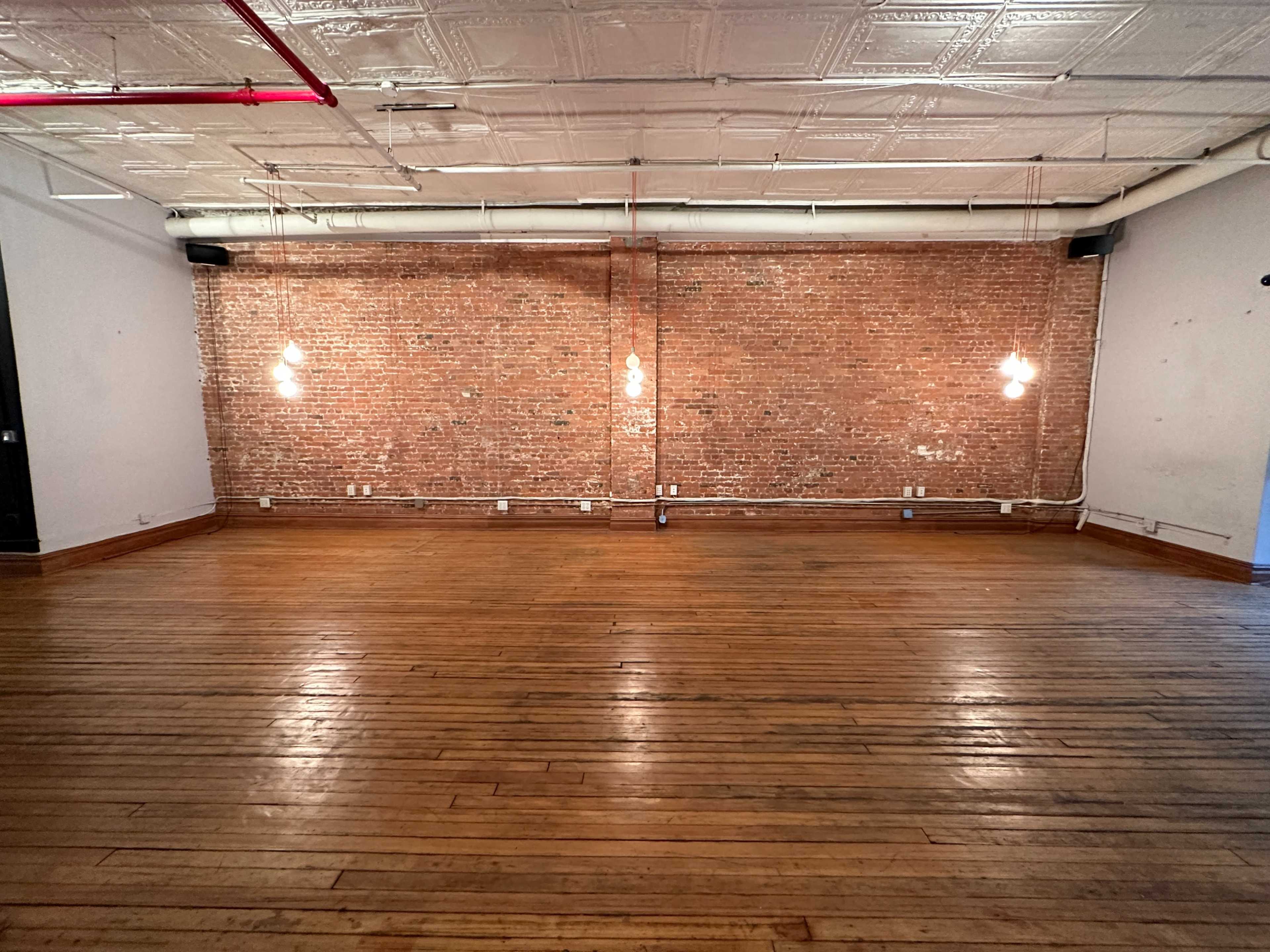 An empty room with exposed brick walls, wooden flooring, and three pendant lights hanging from the ceiling.