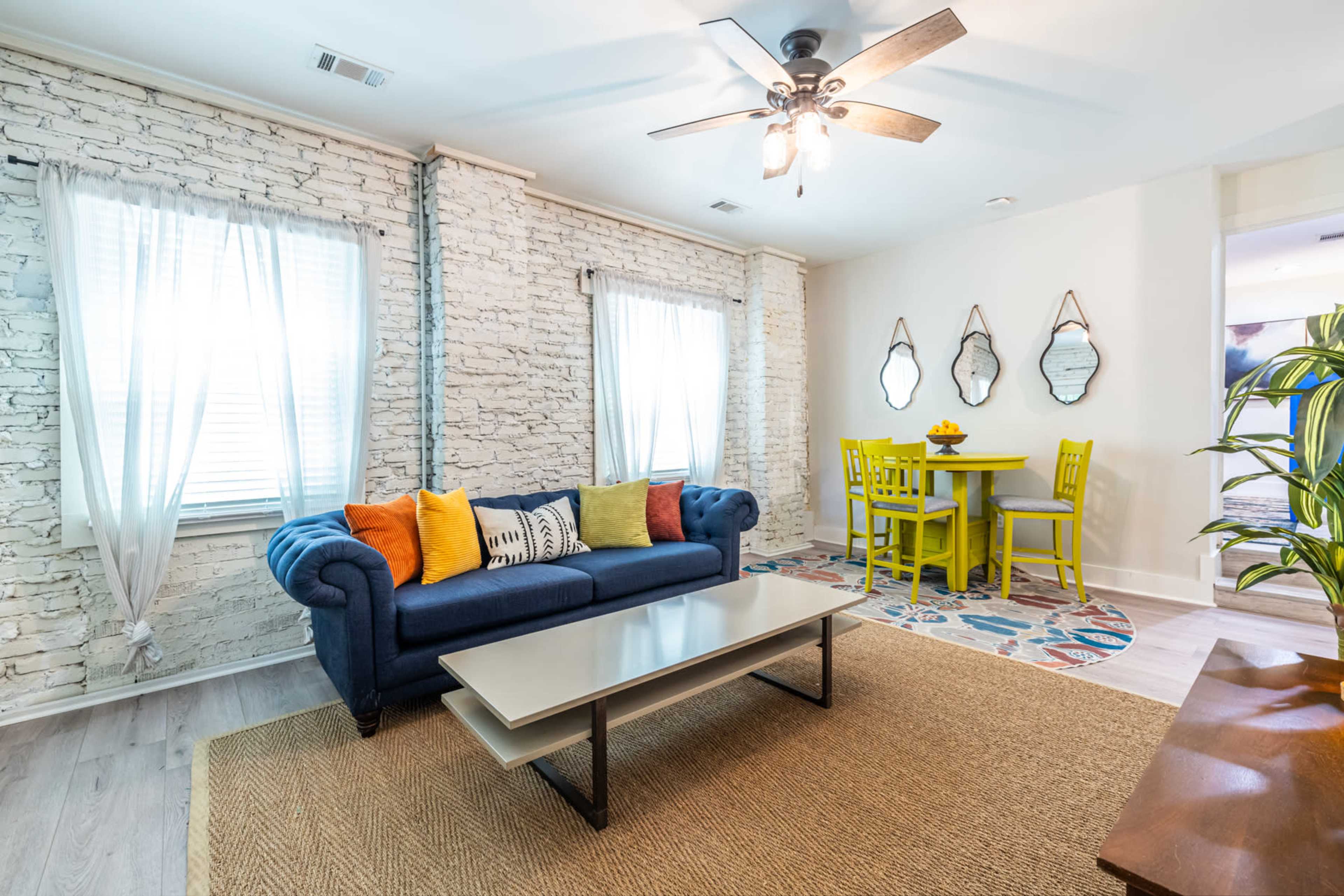 The image shows a cozy living area with a blue couch, a light-colored coffee table, and a small dining table with green chairs, set against a backdrop of white brick walls and large windows.