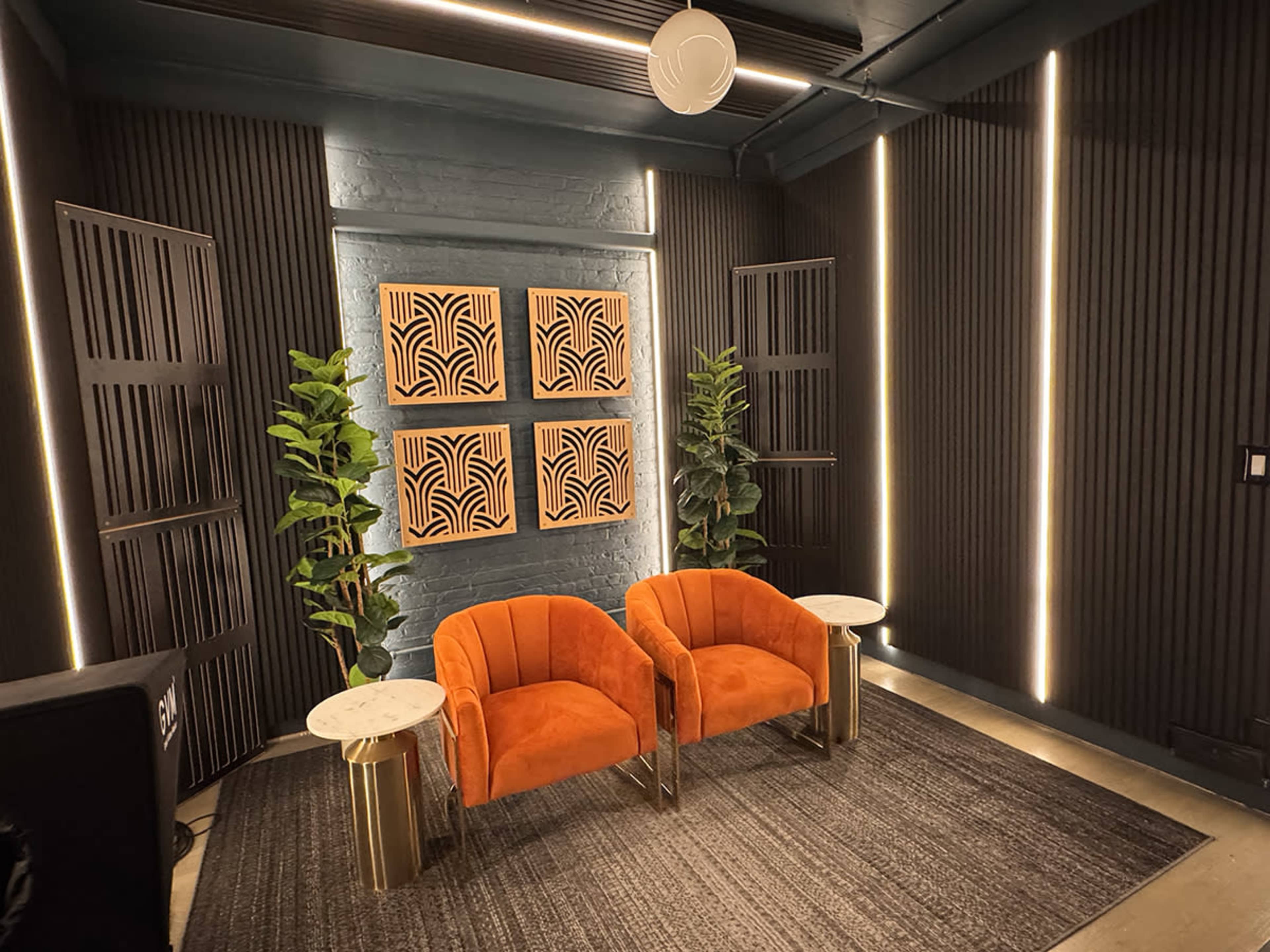 The image shows a modern interior space featuring two orange armchairs, a textured rug, decorative wall panels, and plants, all illuminated by ambient lighting.