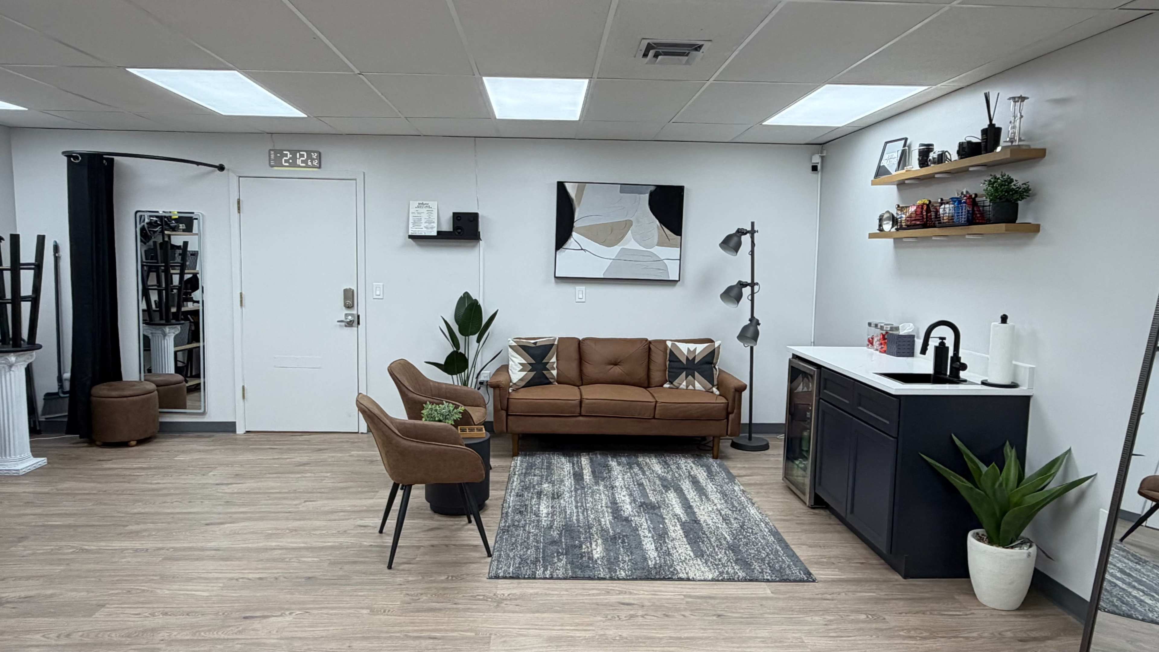 The image shows a modern interior space featuring a brown sofa, a small kitchenette, and decorative plants against white walls and wooden flooring.