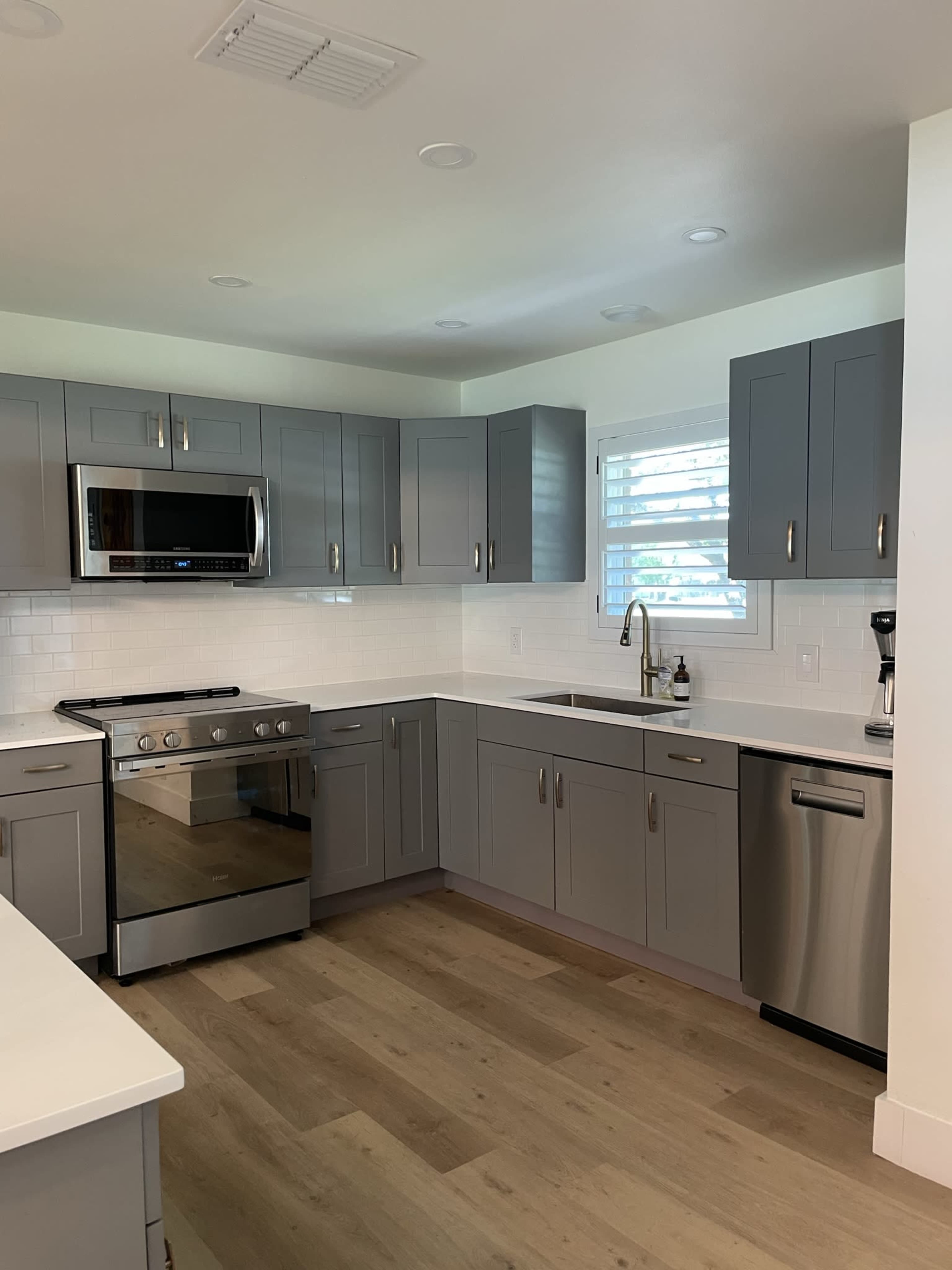 A modern kitchen featuring gray cabinets, stainless steel appliances, and a white countertop with a sink and window.